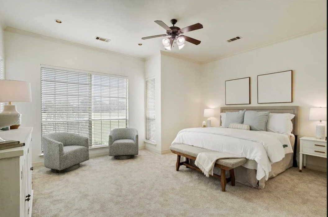 Bright bedroom with large window, light-colored walls, bed with white bedding, two nightstands with lamps, two framed pictures above the bed, two gray armchairs near the window, ceiling fan with lights, carpeted floor.