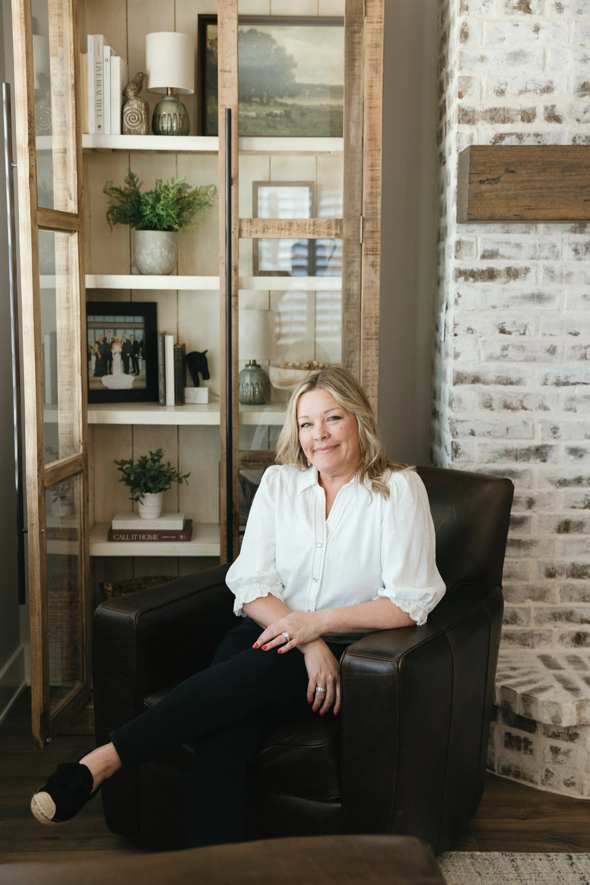 Designer and home stager business owner sitting in a dark brown leather chair in a cozy living room, smiling at the camera. Behind her is a wooden bookshelf with decorative items, plants, and framed photos, and a white brick wall. Feels like home.