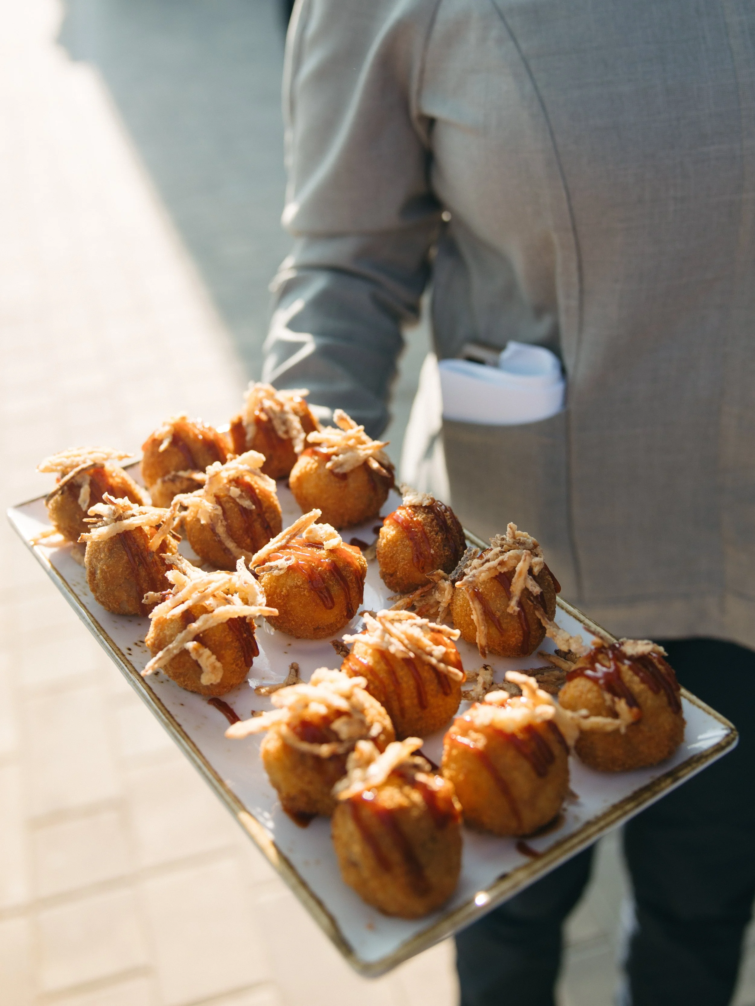 A person holding a rectangular tray filled with fried appetizers garnished with shredded cheese and drizzled with sauce.
