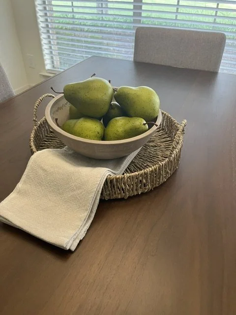 A bowl of green pears on a woven tray, with a white cloth napkin underneath, on a dark wooden dining table near a window with blinds.