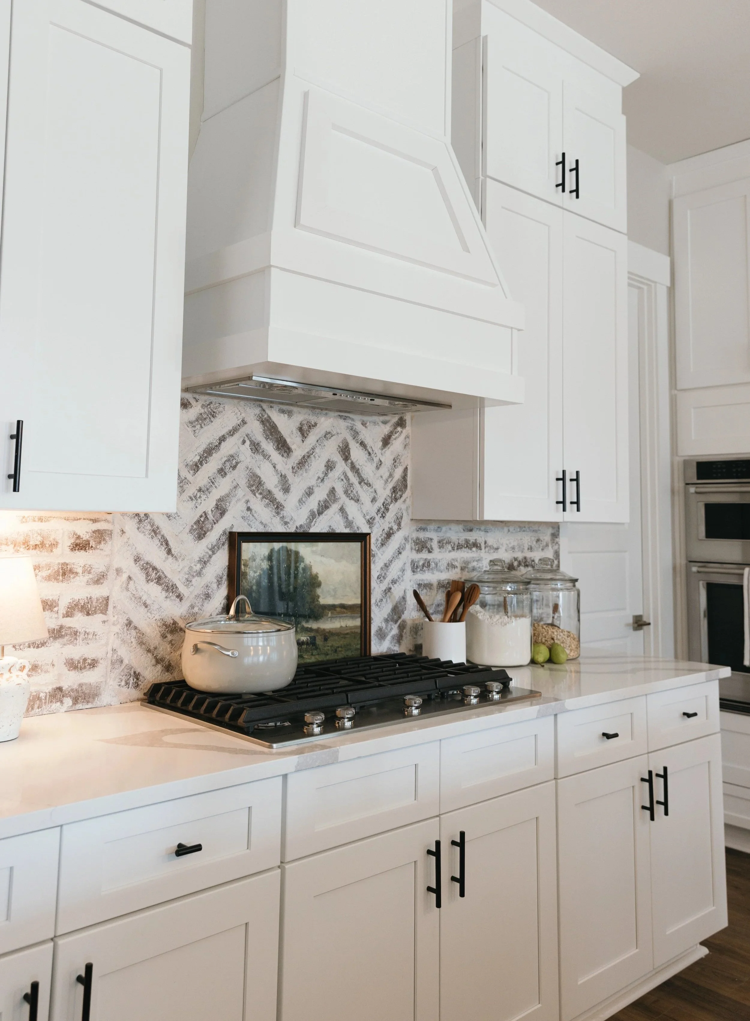 White kitchen with white cabinets, a stovetop with a white pot, decorative jars, a framed landscape picture, and a textured brick backsplash.