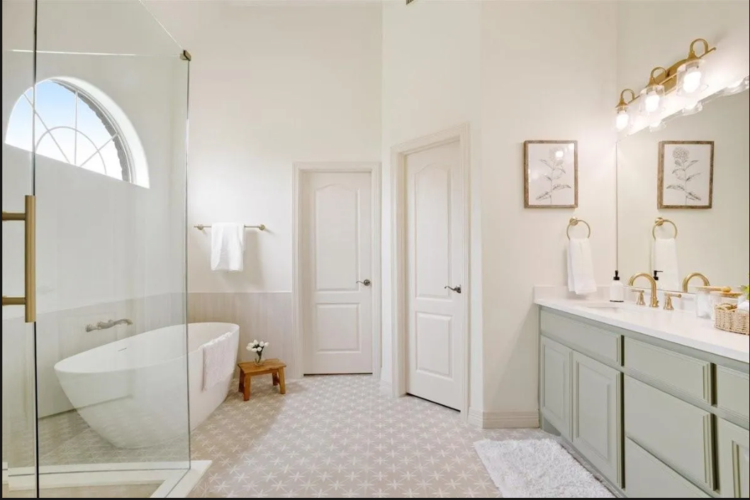 Bright bathroom with a glass-enclosed shower, free-standing bathtub, double vanity with two sinks, framed artwork, and light fixtures with multiple bulbs.