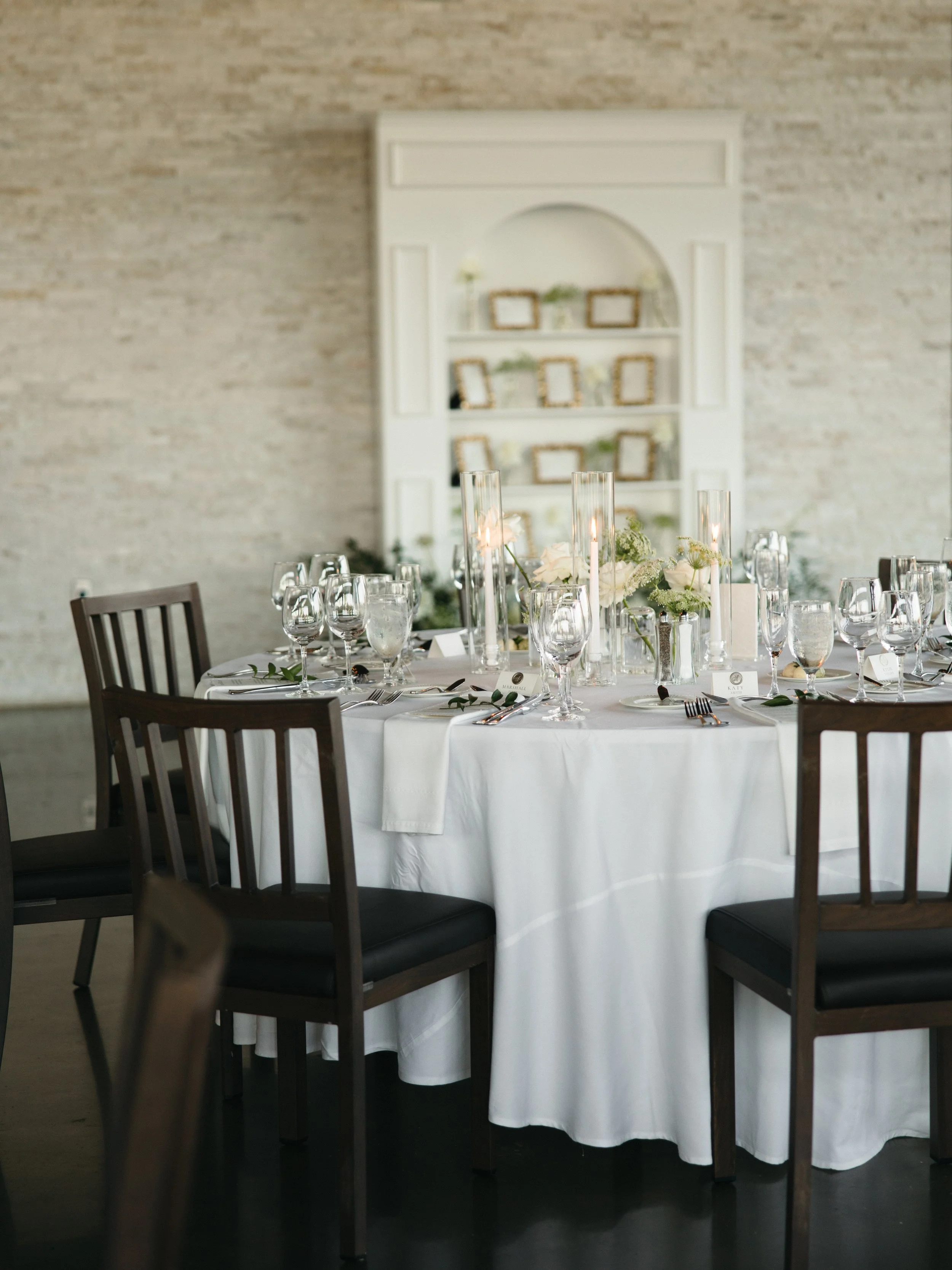 Elegant round table with white tablecloth, set for a formal event with multiple wine glasses, silverware, a centerpiece with candles and flowers, surrounded by dark wooden chairs, in a room with a textured beige wall and a white bookshelf with picture frames and decorative items in the background.