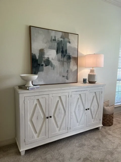 A white wooden sideboard with four cabinet doors, each featuring a diamond pattern, placed against a beige wall. On top of the sideboard, there is a white bowl, some books, a table lamp with a pink shade, a small candle, and a decorative vase. Above 