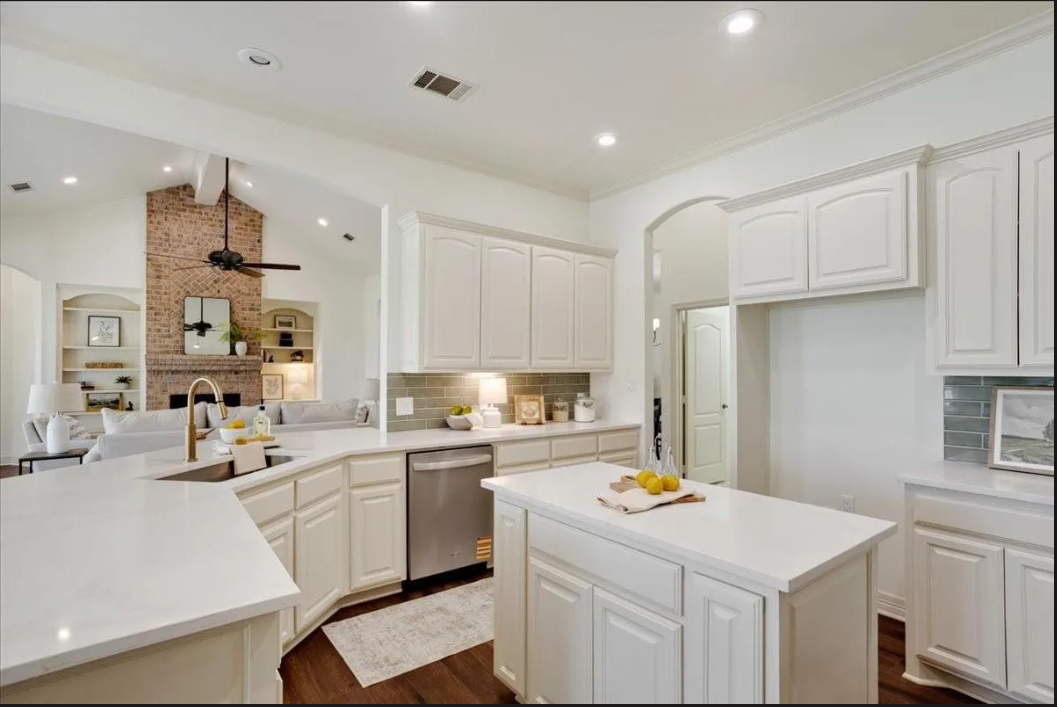 Modern kitchen with white cabinets, a kitchen island with a lemon on a towel, stainless steel dishwasher, gray backsplash, and open to a living room with a brick fireplace and ceiling fans.