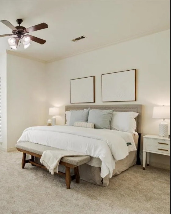 A bedroom with a neatly made bed, framed artwork above the headboard, two bedside tables with lamps, a ceiling fan, and neutral-colored decor.