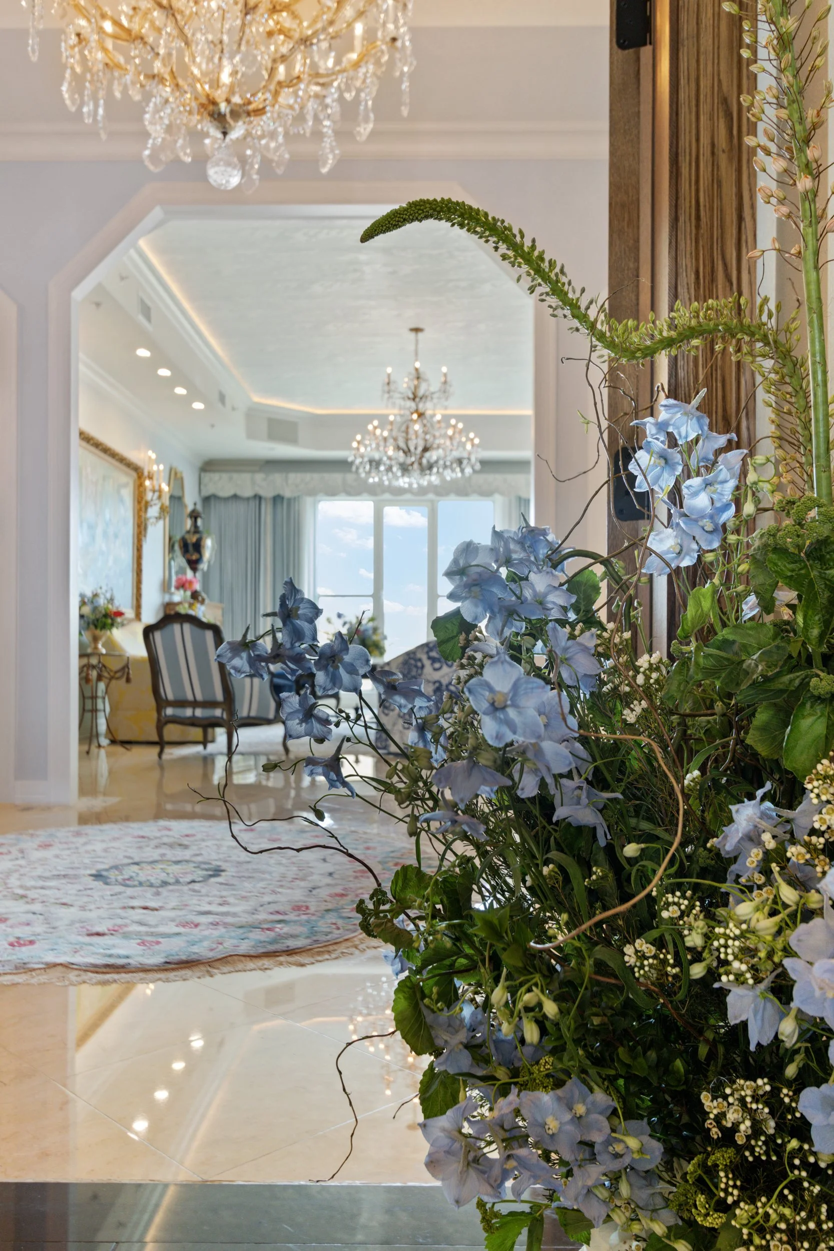 View of a luxurious living room with a large flower arrangement in the foreground, featuring blue flowers and green foliage. The room includes elegant chandeliers, a patterned area rug, a marble floor, French doors with drapes, and classic furniture with ornate frames.