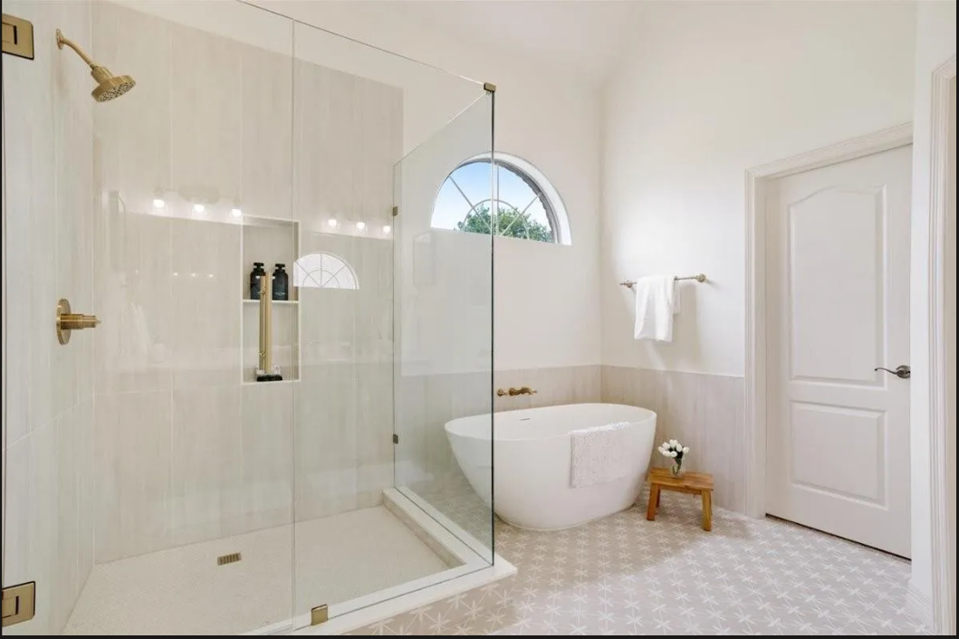 Bright bathroom with a glass-enclosed shower, a freestanding bathtub, a small wooden stool with a flower vase, a white towel, and a window with an arched top.