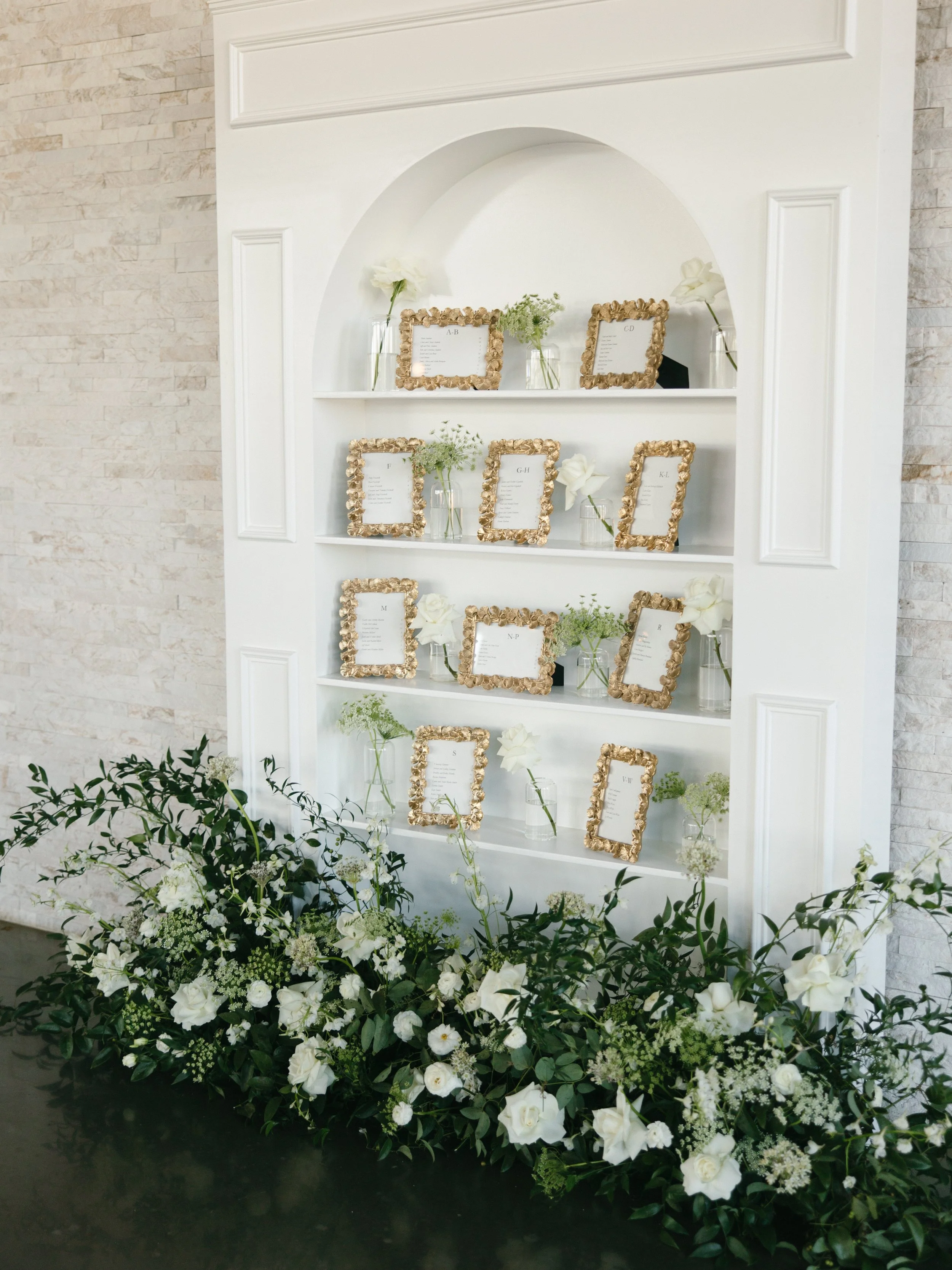 A wedding or event seating chart display with framed place cards on white shelves, decorated with flowers in glass vases, surrounded by a large floral arrangement of white flowers and greenery at the base.