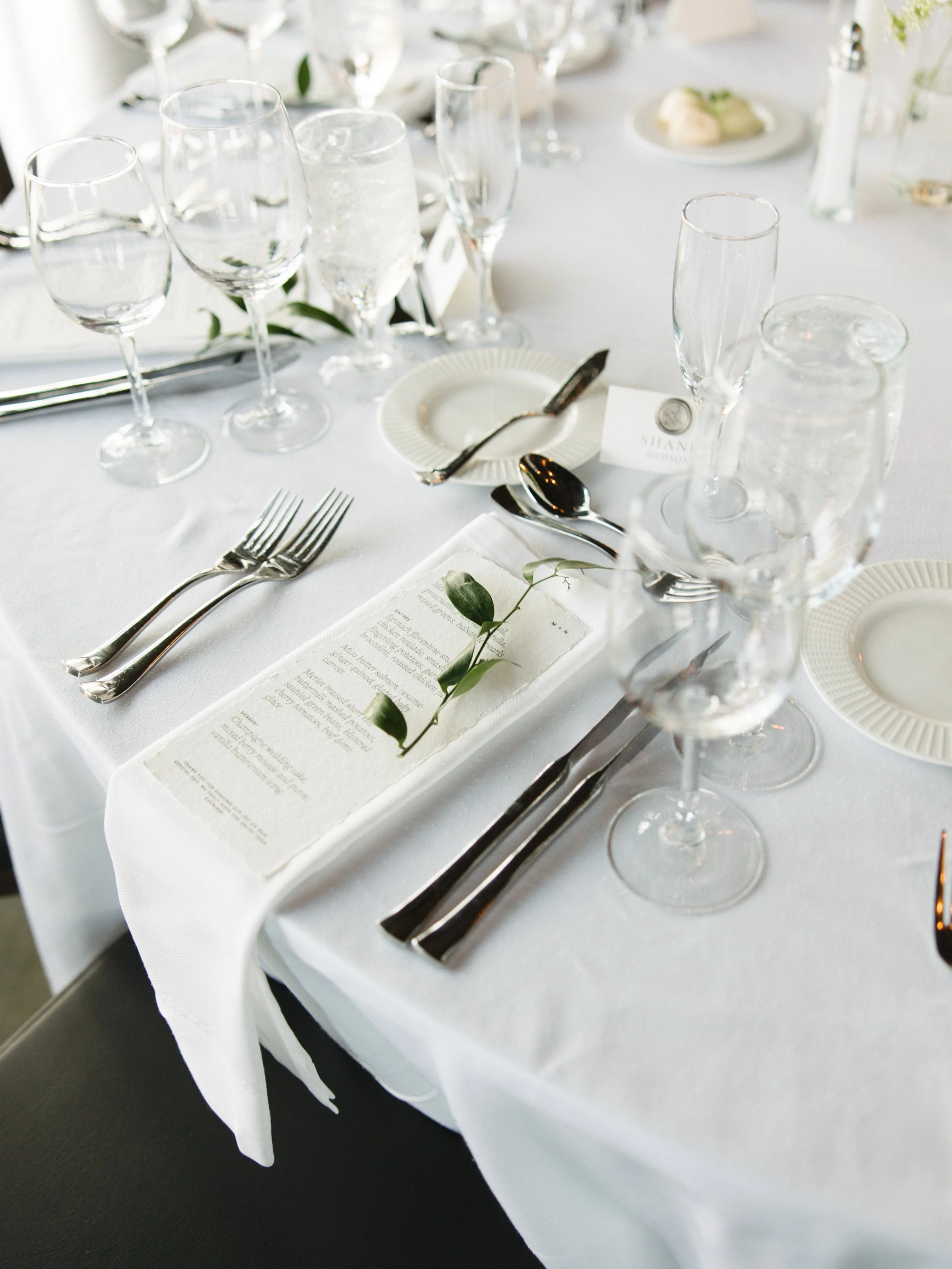 Elegant dining table with multiple wine glasses, water glasses, silverware, a small white plate, a menu, and a place card on a white tablecloth, decorated with greenery.