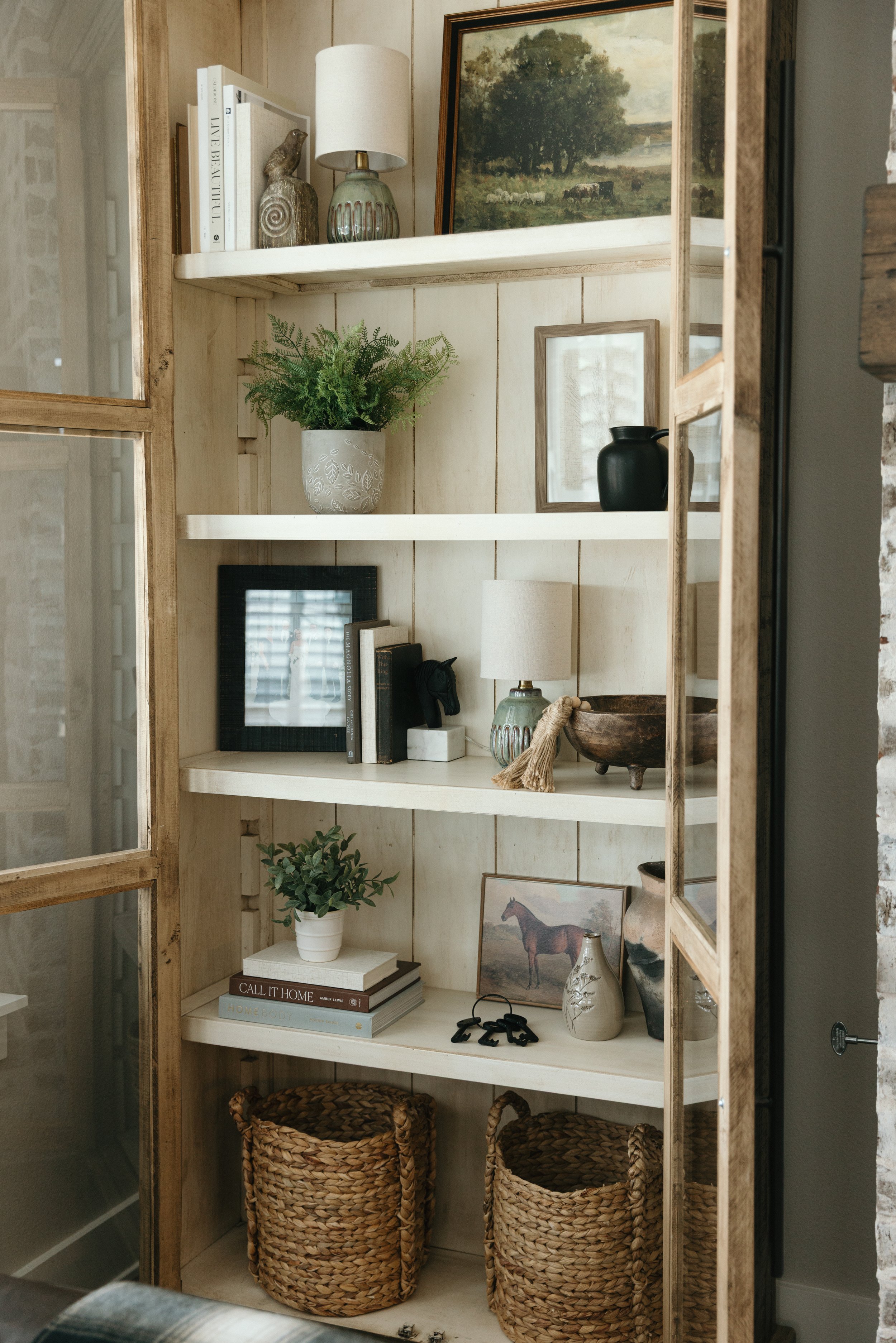 A wooden and white open shelving unit with decorative items including books, vases, framed artwork, a potted plant, and woven baskets.