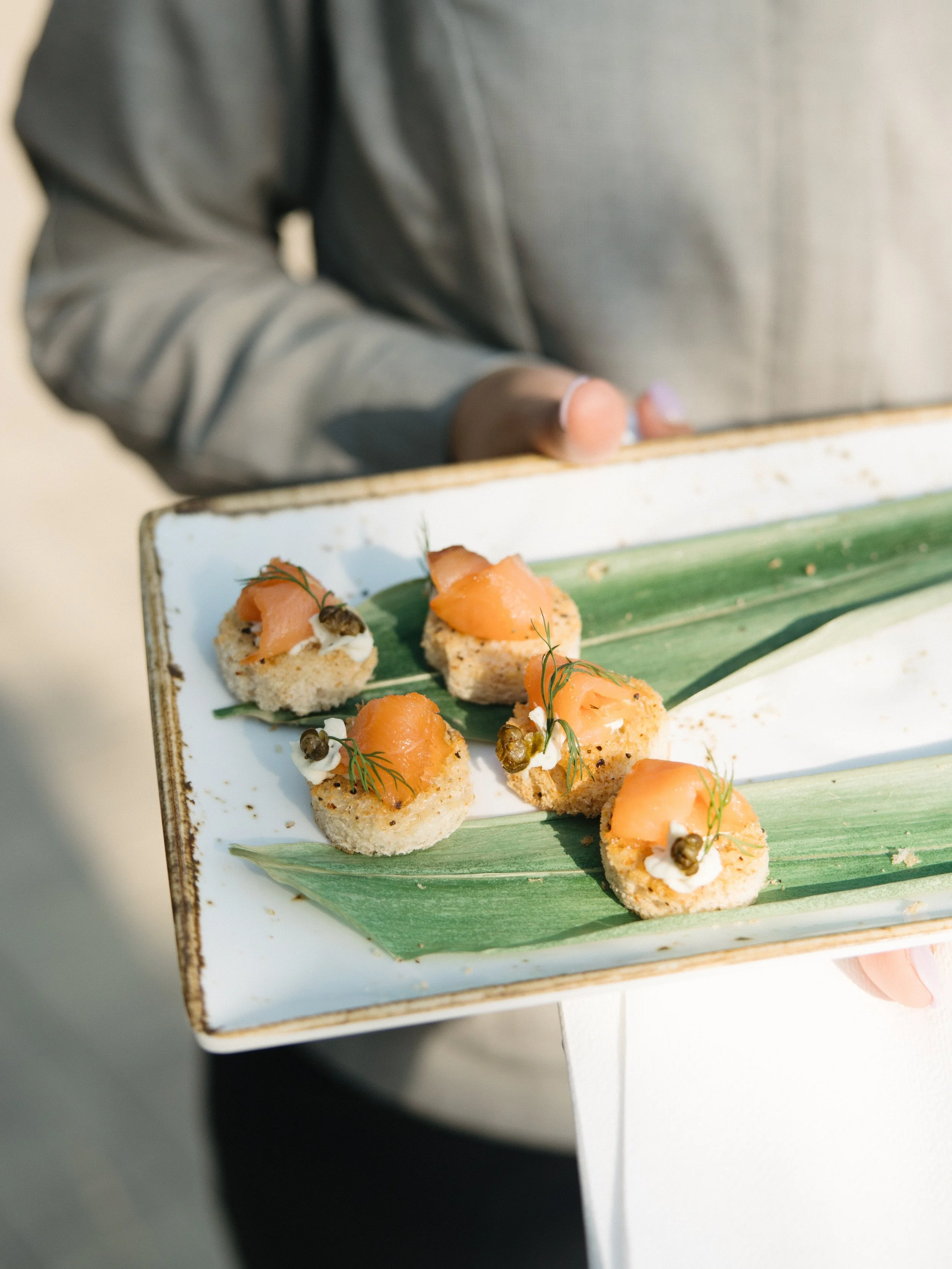 Appetizers with smoked salmon on small toasted bread slices, garnished with herbs, on a rectangular white platter with a green leaf and leaf-shaped decoration.
