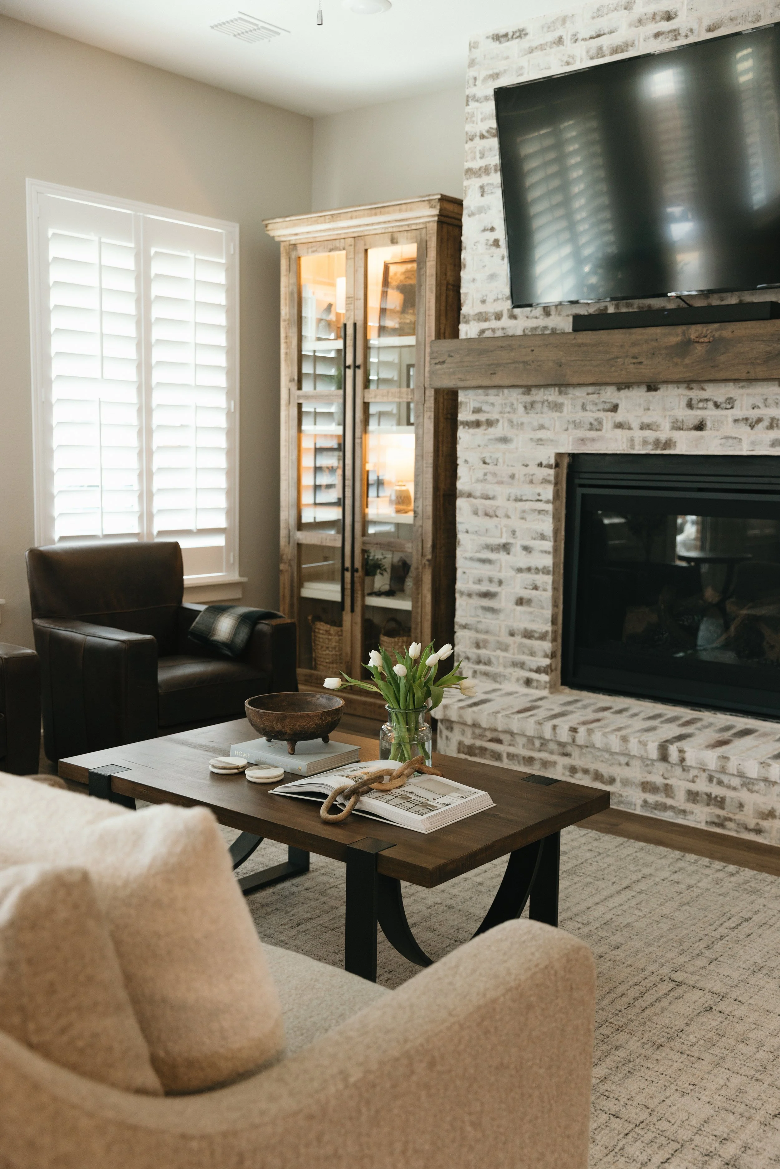 Living room with beige armchair, dark wooden table with a vase of white tulips, open magazines, and decorative items; fireplace with white brick surround and dark wooden mantel; wall-mounted TV; wooden cabinet with glass doors; large window with white plantation shutters.