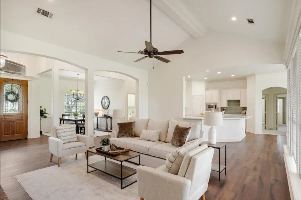 Bright living room with white walls, ceiling fan, beige sofas, armchairs, black coffee table, dining area, and white kitchen in the background.