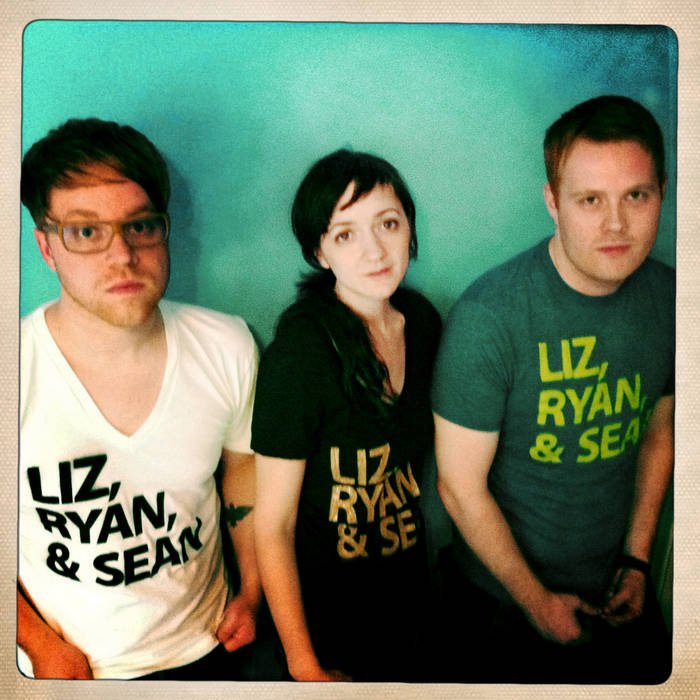 Three people sitting against a green wall, all wearing T-shirts with the text "Liz, Ryan, & Sean."