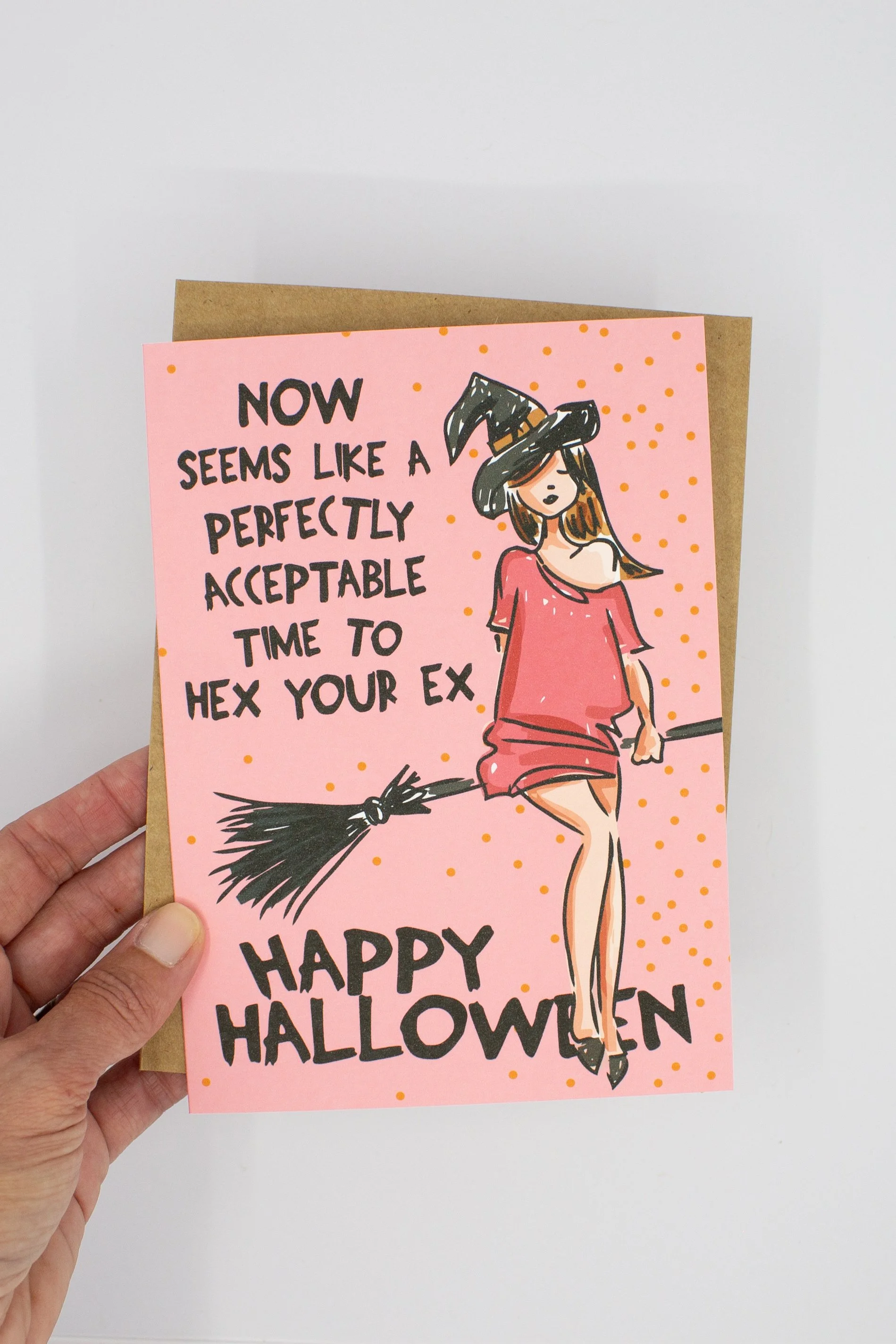 Halloween greeting card featuring a cute witch on her broom against a playful pink background with orange dots.