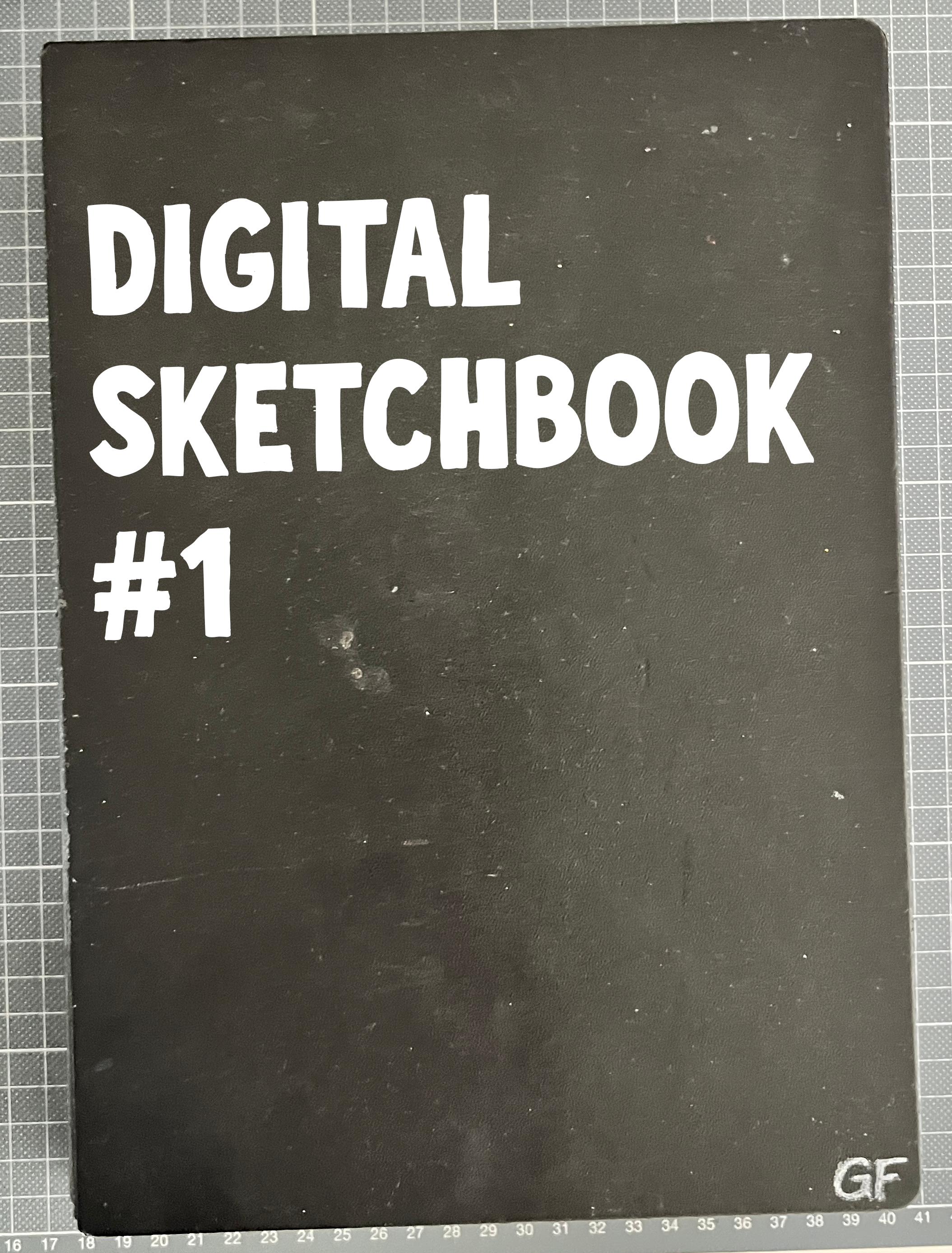 Black sketchbook cover hand-lettered 'Digital Sketchbook #1' with a 'GF' monogram, resting on a gridded cutting mat