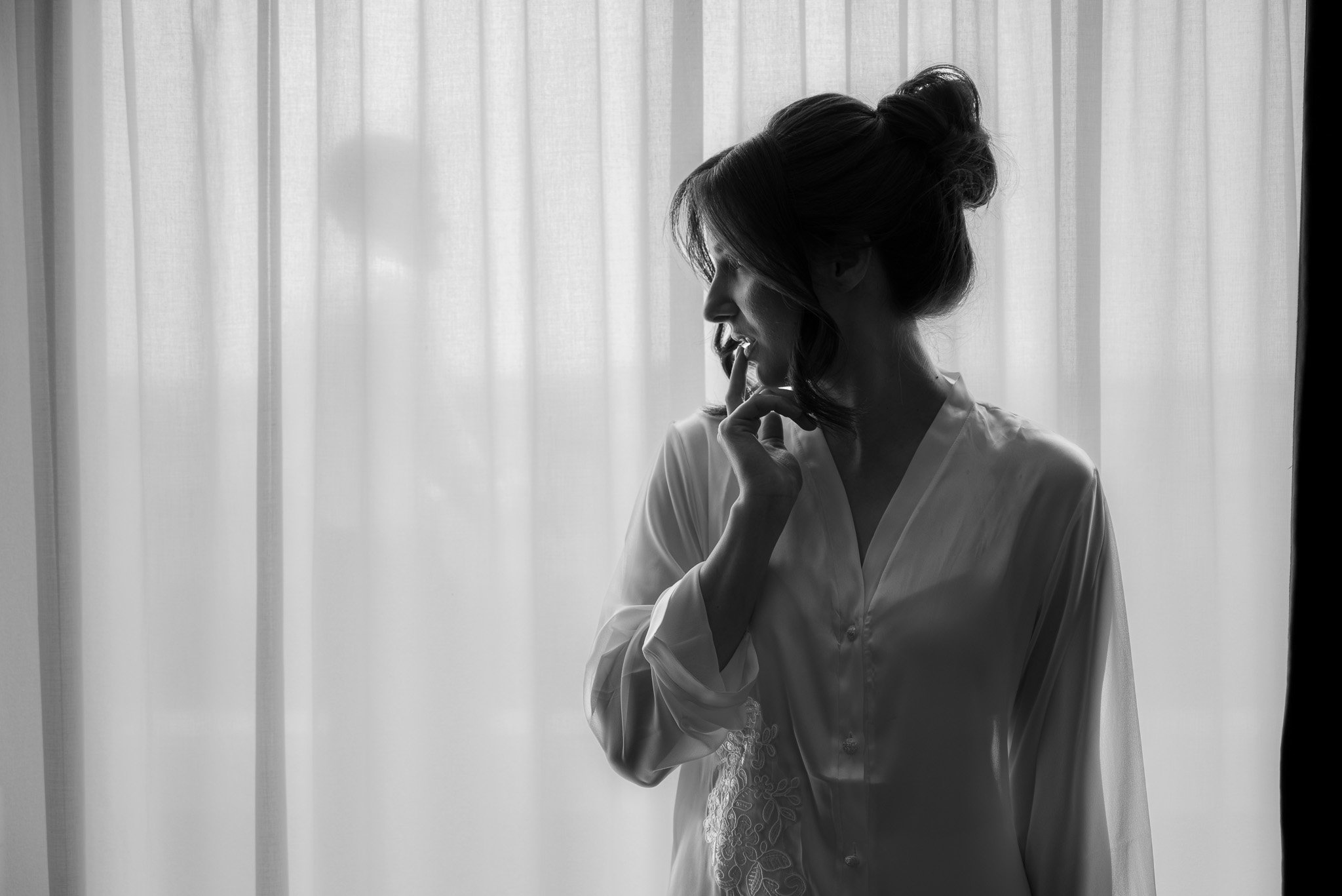 Black and white photo of a woman in profile with her hand near her mouth, standing in front of a sheer curtain.