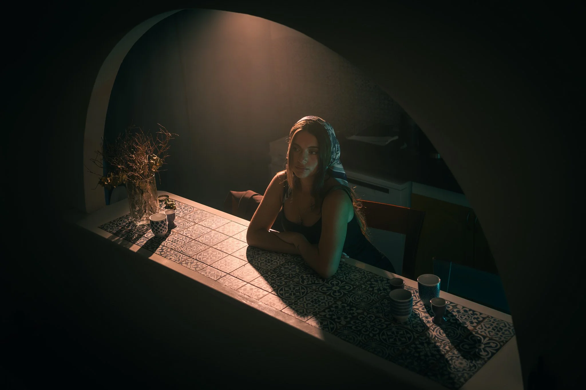 Fotografie ženy s dlouhými tmavými vlasy, v černém topu a šátku na hlavě, sedící u stolu se vzorovanými dlaždicemi v tlumeně osvětlené místnosti s měkkým světlem svítícím shora, s vázami a šálky na stole.
