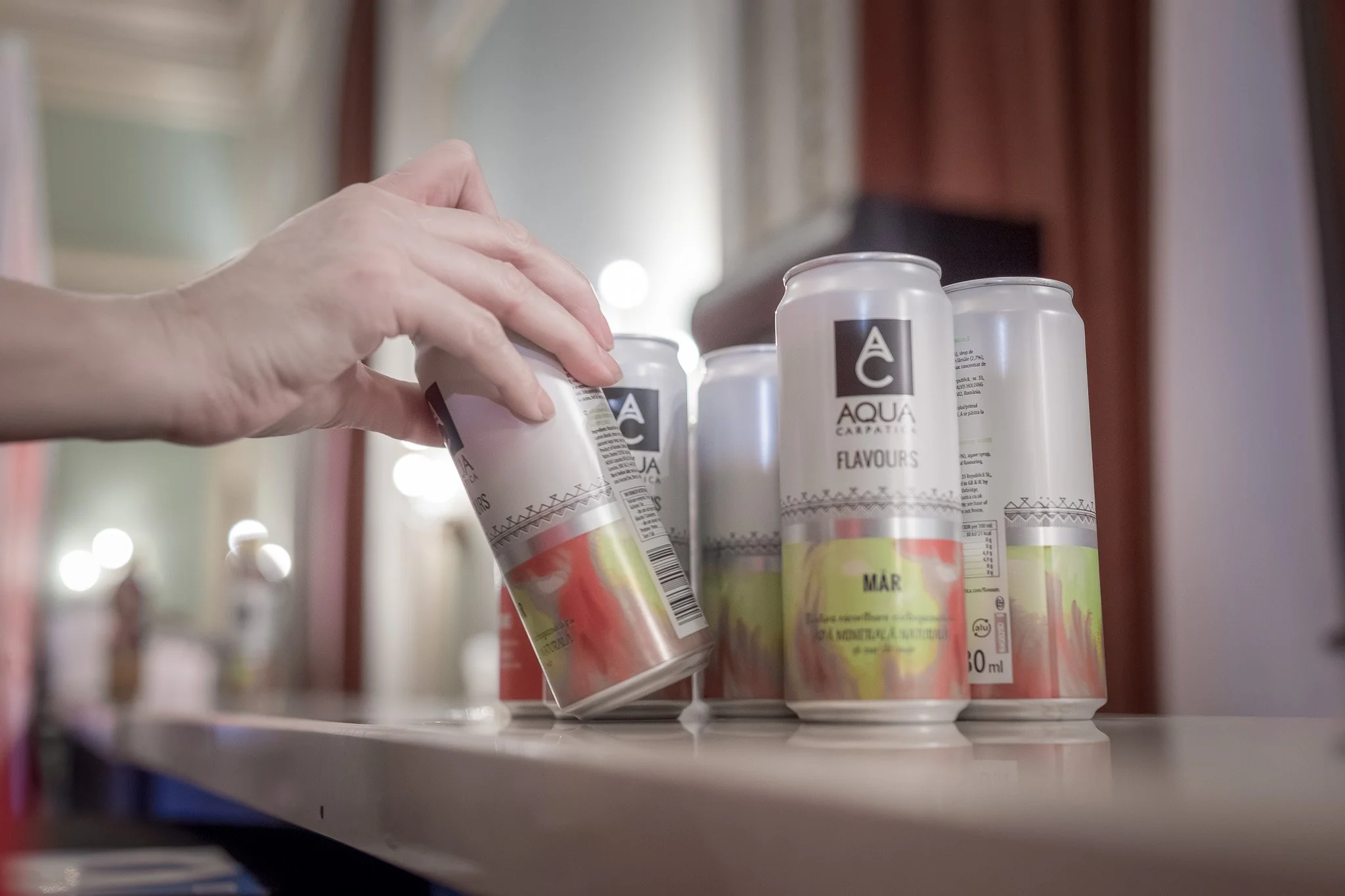 A hand placing a can in a row of identical cans with white backgrounds and black and green labels, on a beige surface inside a room with curtains and windows in the background.