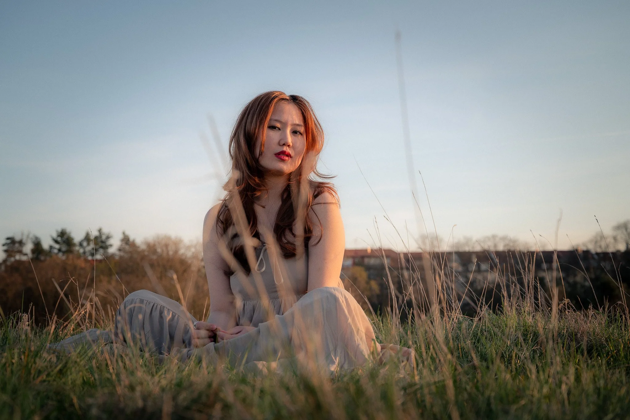 A woman with long brown hair sitting cross-legged in a grassy field during sunset, wearing a beige dress.