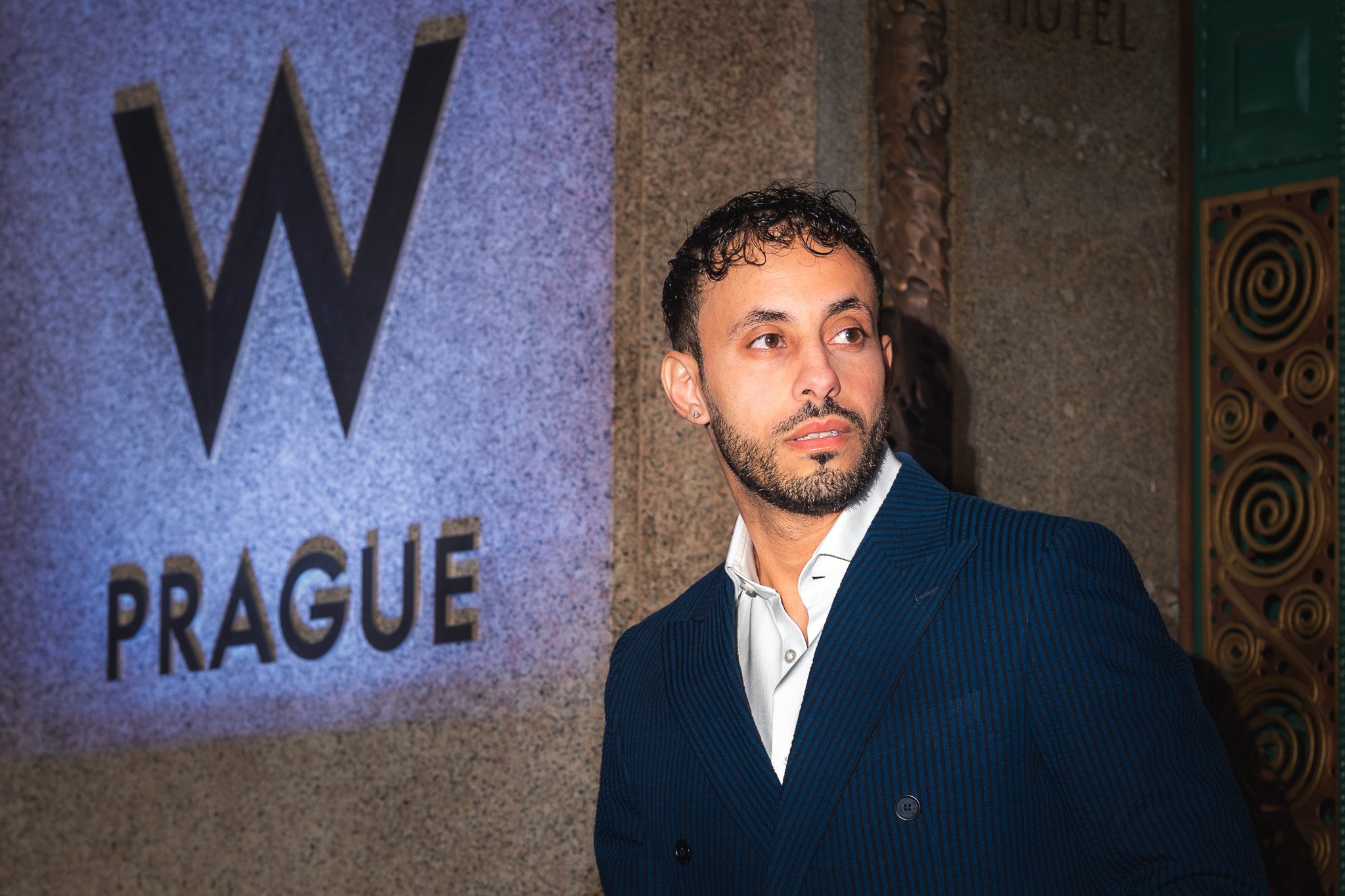 Muž v modrém pruhovaném obleku stojí u vstupu s nápisem „W Prague“ a dívá se do strany; kamenná stěna a dekorativní kovové dveře.