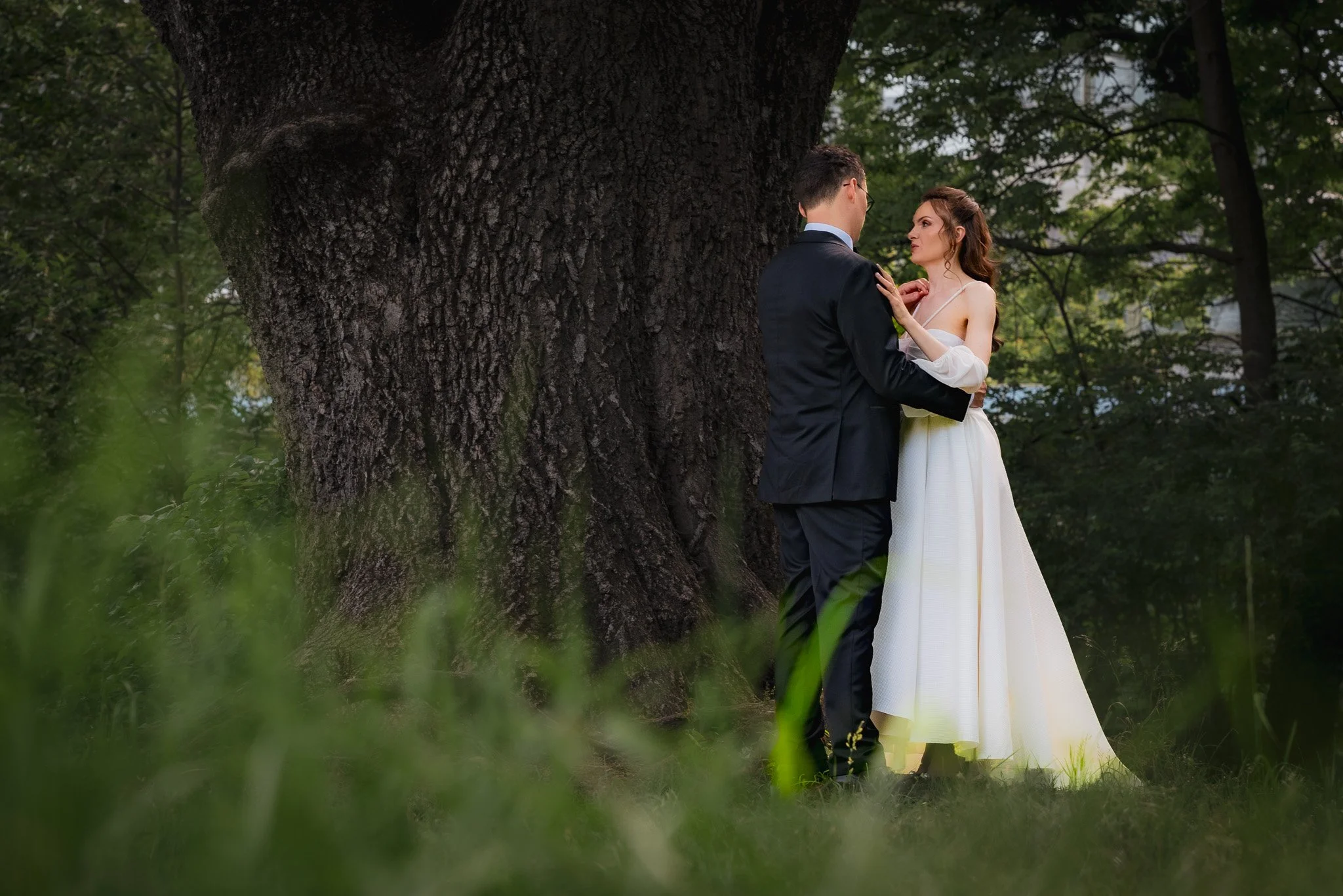 A bride and groom standing close together outdoors near a large tree, gazing into each other's eyes. The bride is wearing a white strapless wedding dress, and the groom is in a black suit. The scene is set in a green forest.