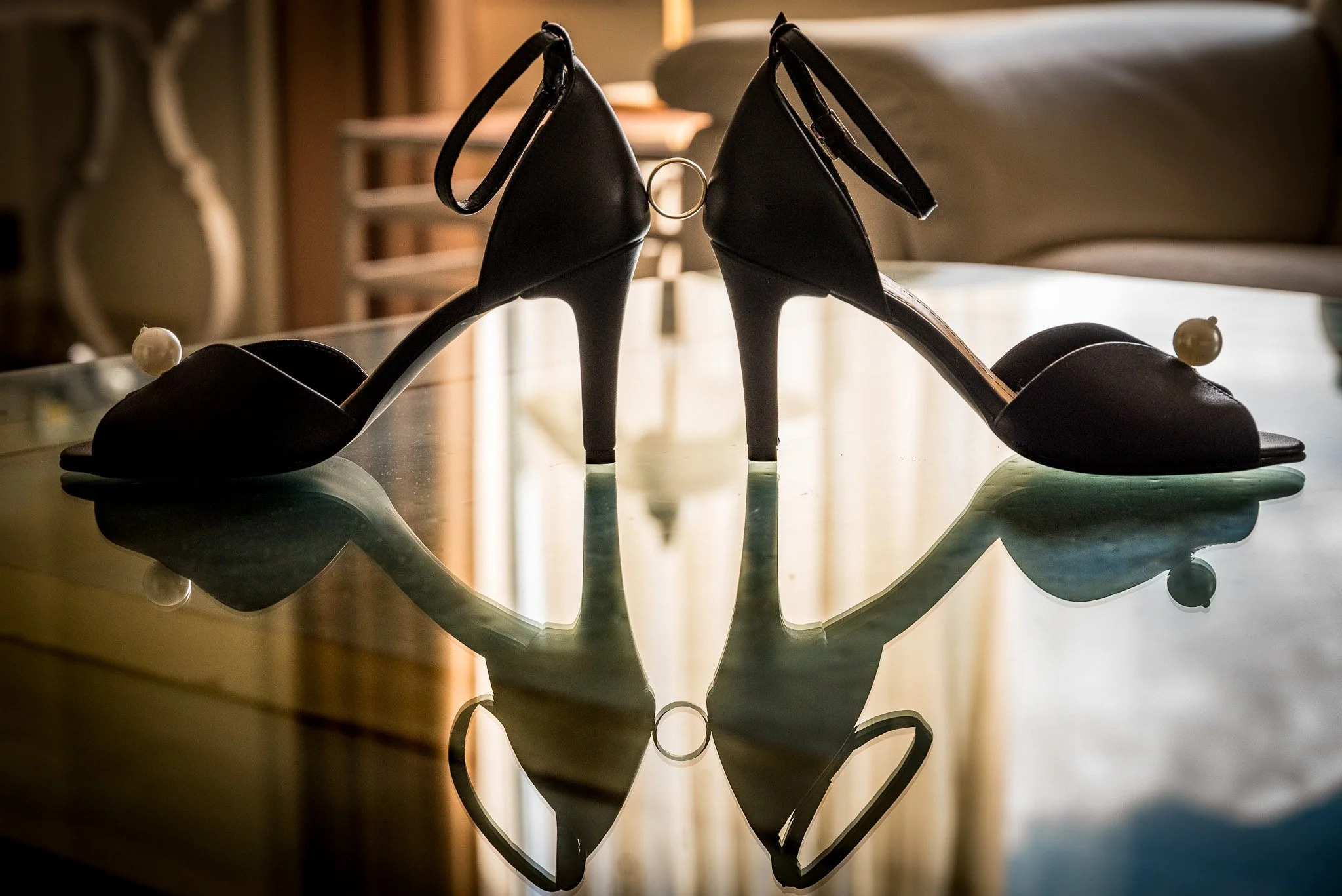 Black high-heeled shoes with ankle straps and pearl details on a reflective glass surface.