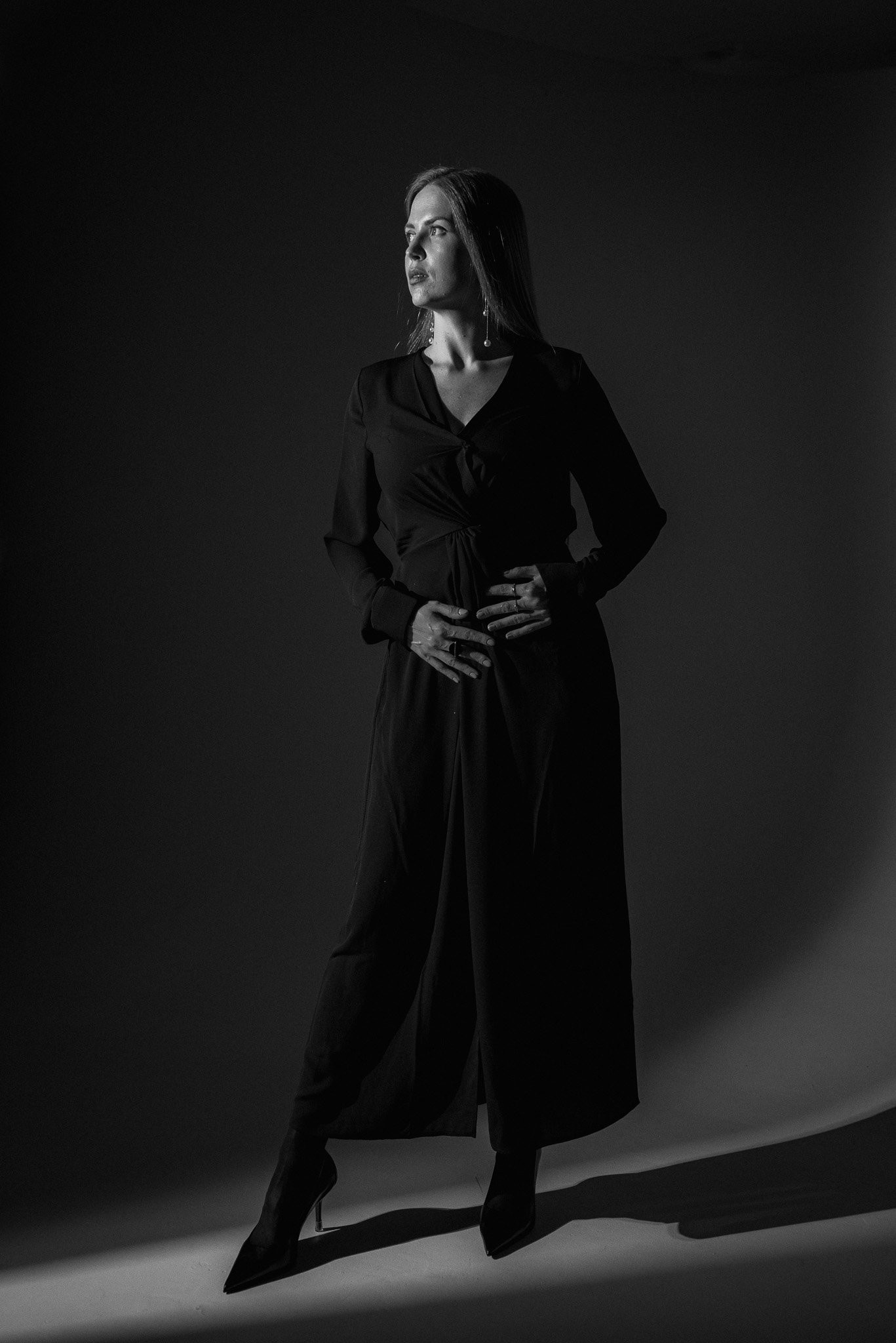 Black and white portrait of a woman in a long dress and heels, posing dramatically with side lighting highlighting her profile.
