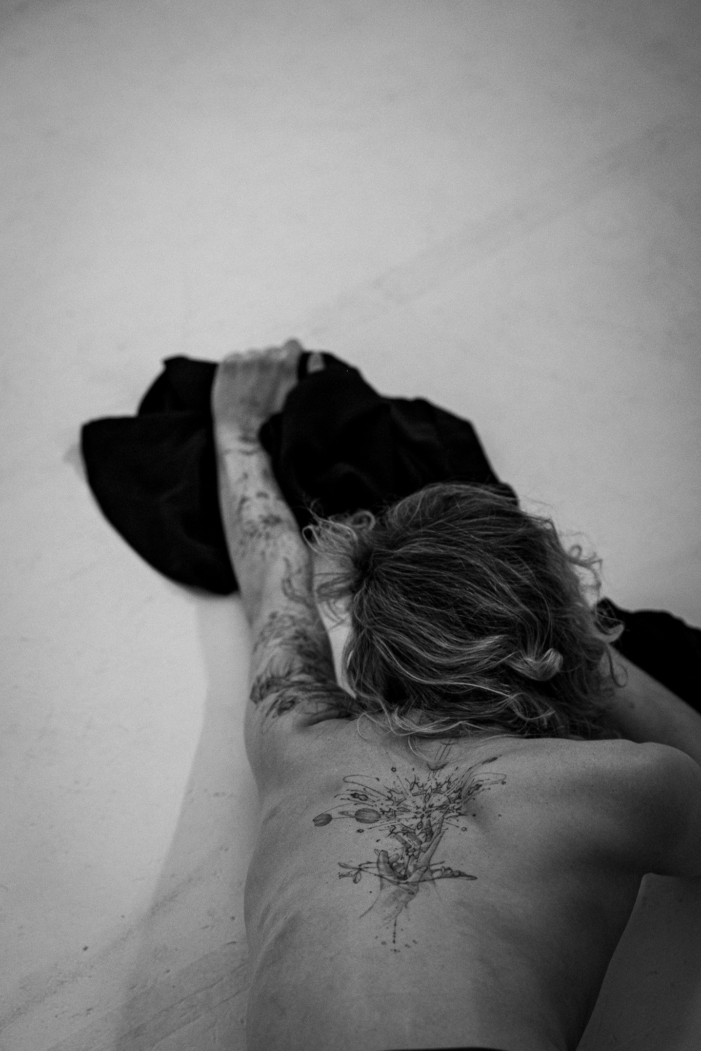 Black and white image of a person with a back tattoo, lying face down on a surface with an outstretched arm holding a dark cloth.