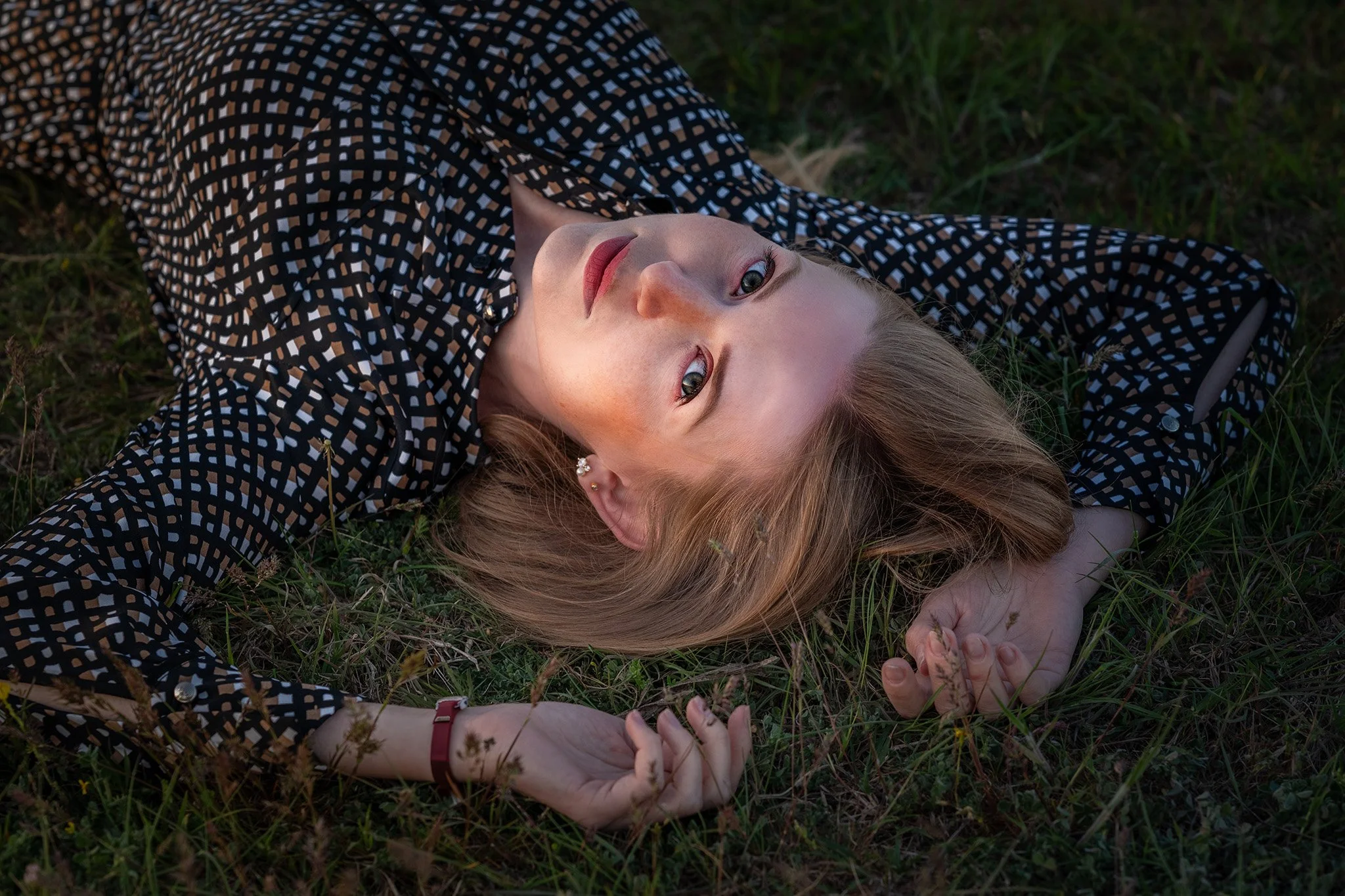 A young woman with blonde hair lying on grass, looking up at the camera, wearing a black and white patterned top, with her hands resting near her head.