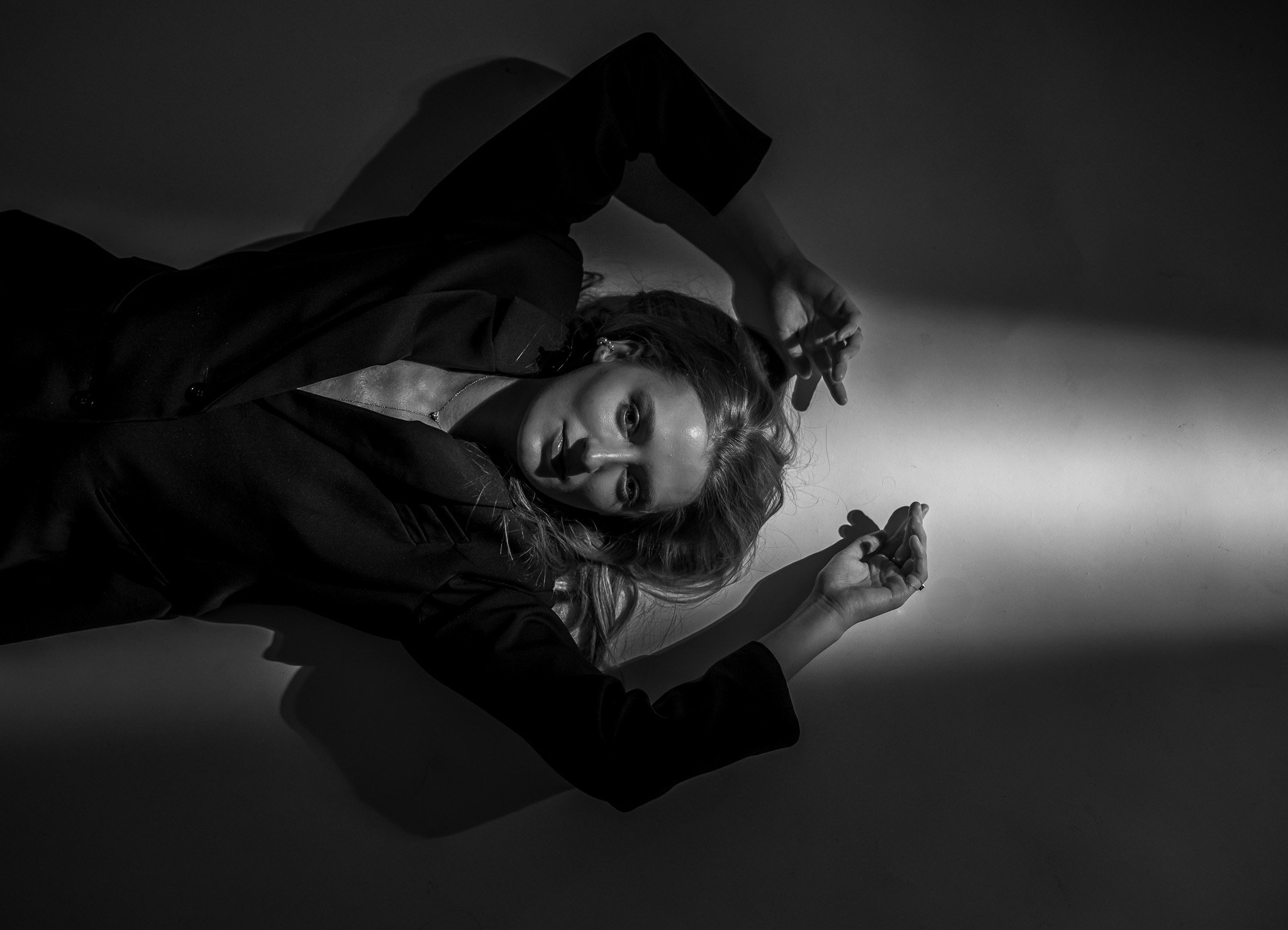 Black and white photo of a person in a black outfit lying on the floor with arms above their head, dramatic lighting casting shadows.