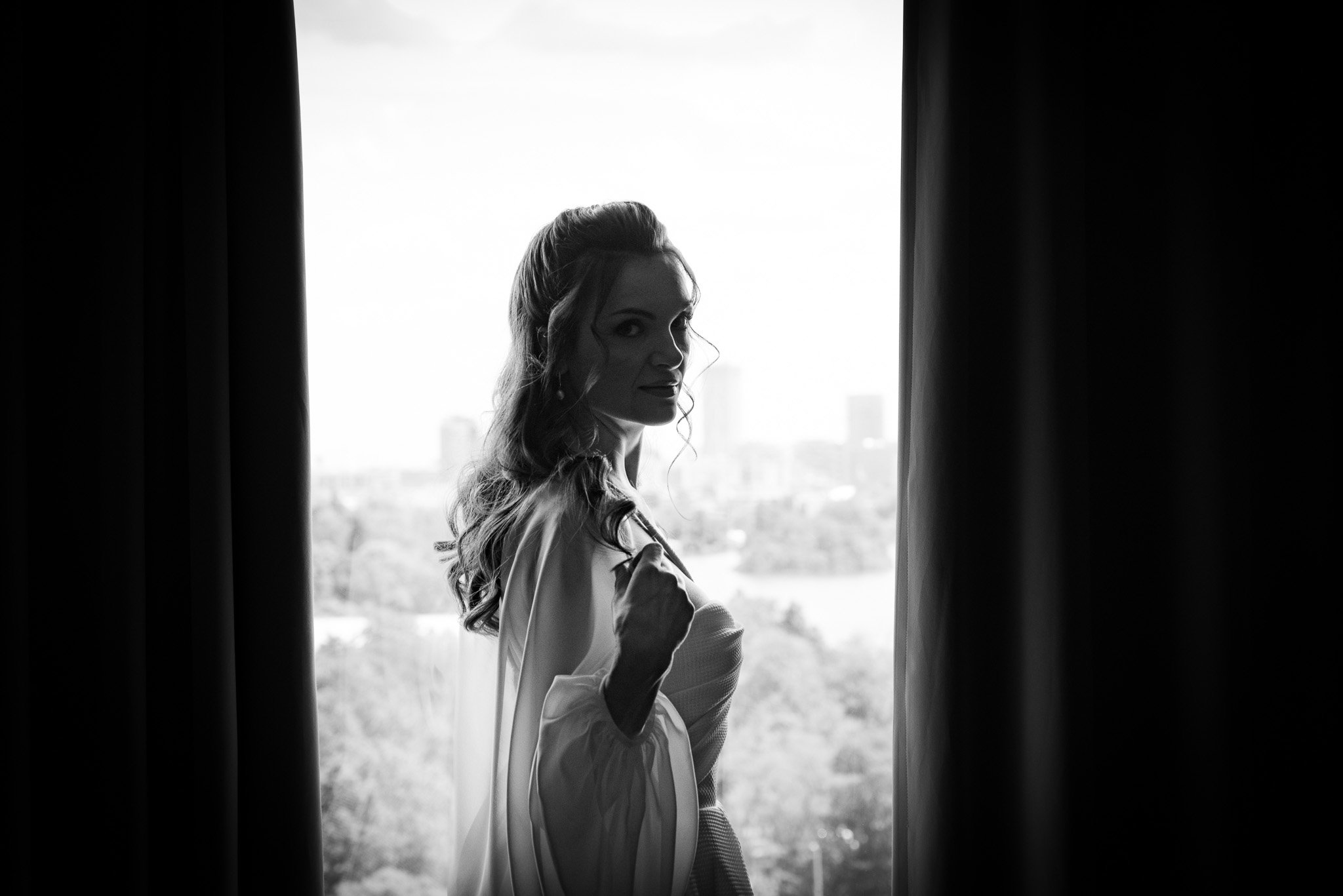 Silhouette of a woman with long hair standing between curtains, looking outside a window, with a cityscape in the background.