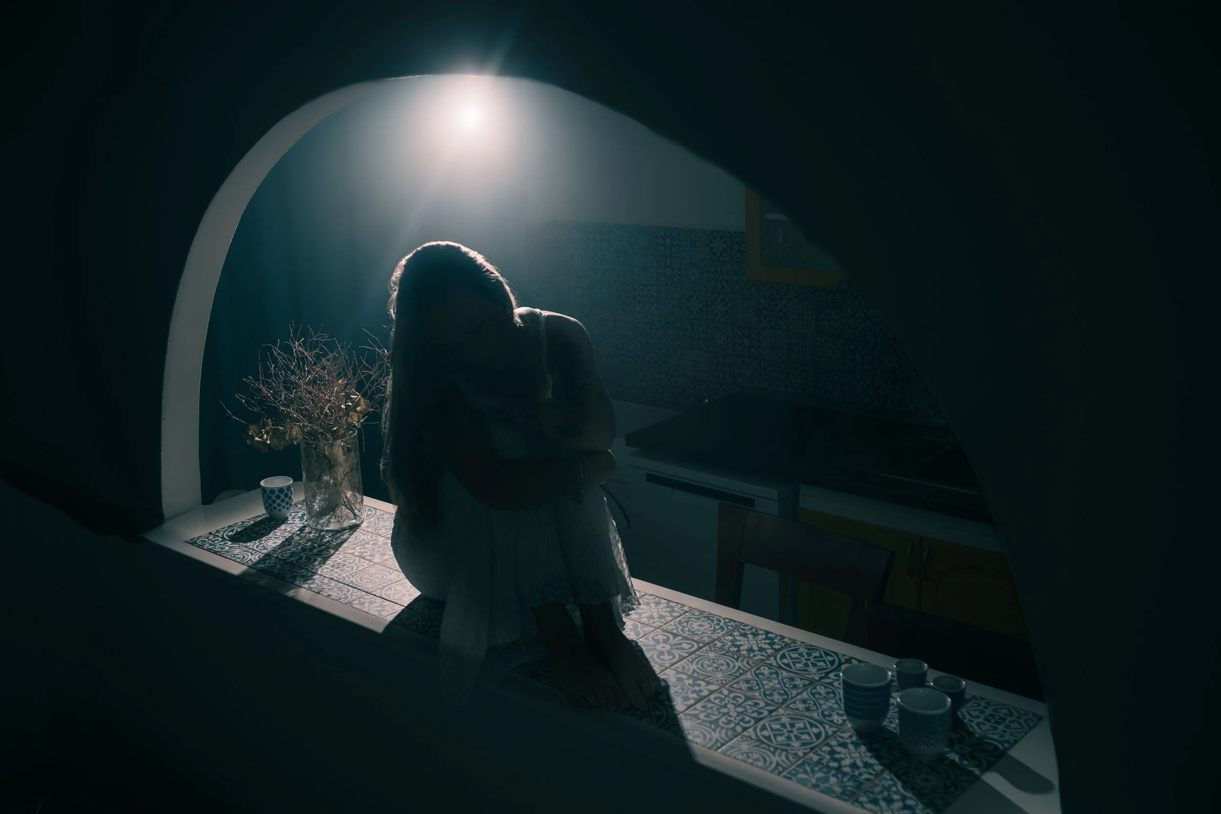 Fotografický portrét mladé dívky s dlouhými vlasy stojí v tlumeně osvětlené místnosti ozářené slunečním světlem, otočená zády k fotoaparátu. Stojí u stolu s vázou, na které jsou sušené větve, šálek a několik šálků uspořádaných na vzorovaném ubrusu.