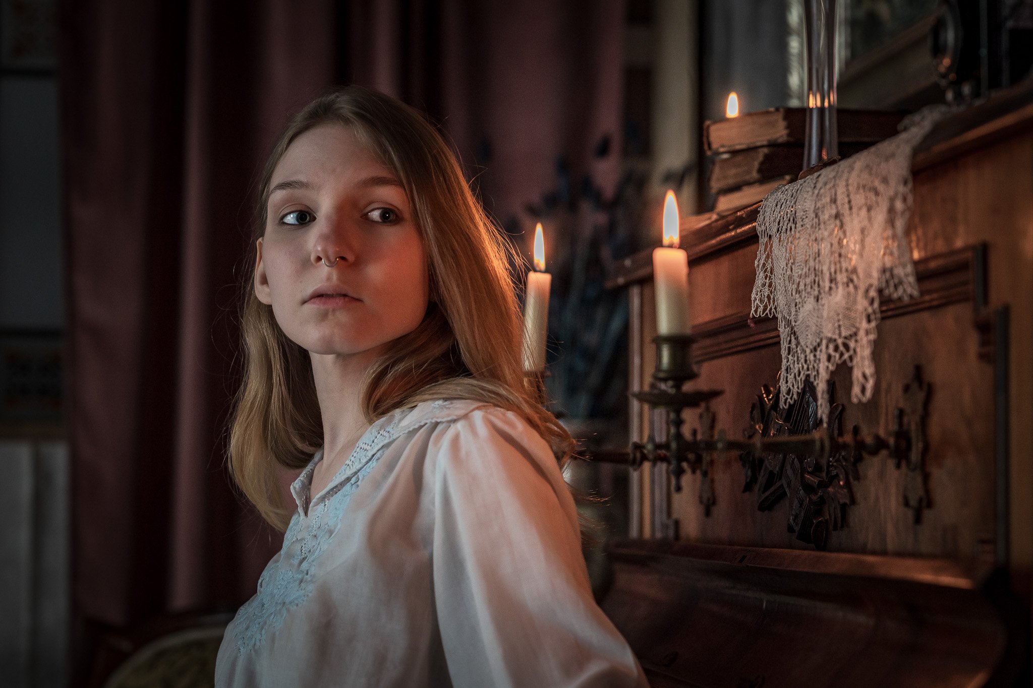 Woman in historical setting by lit candles and lace, wearing a white blouse, looking to the side.