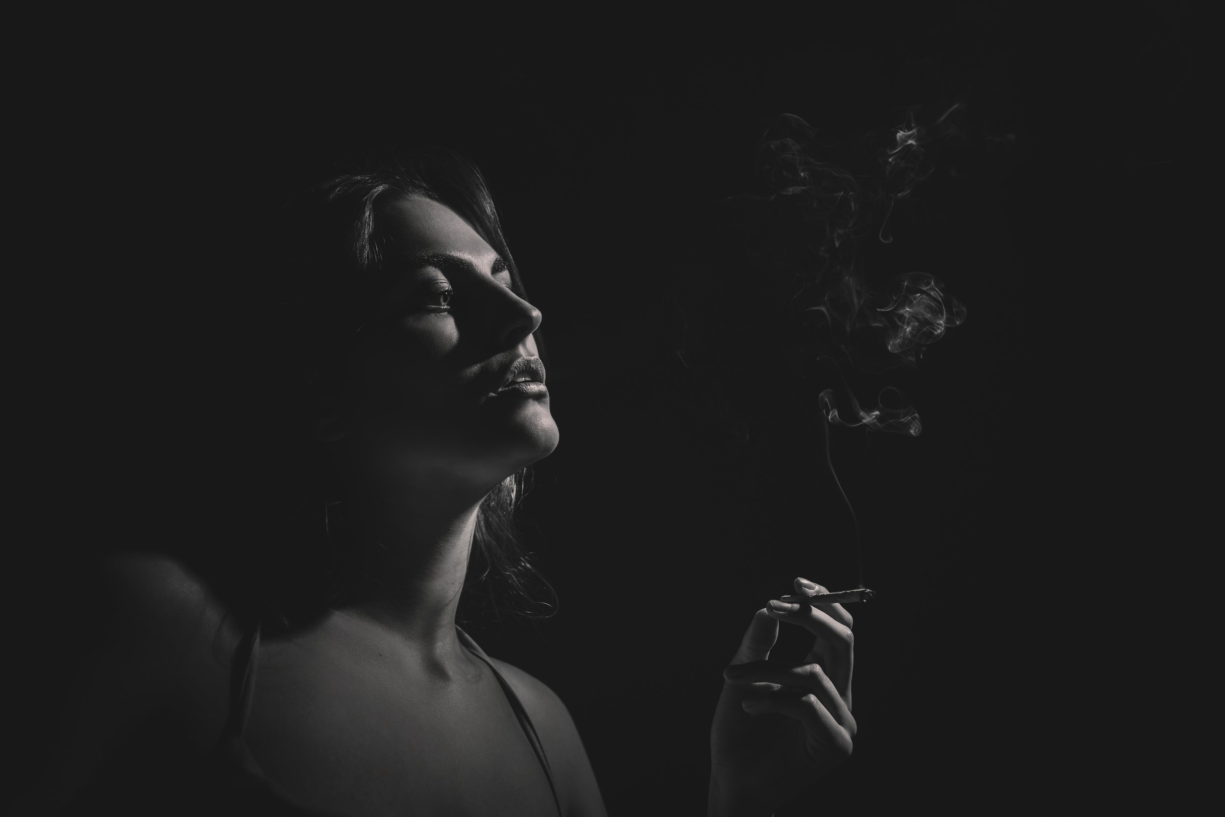 Fotografie ženy v tmavém pražském baru, částečně osvětleném, jak drží cigaretu, z níž stoupá dým.