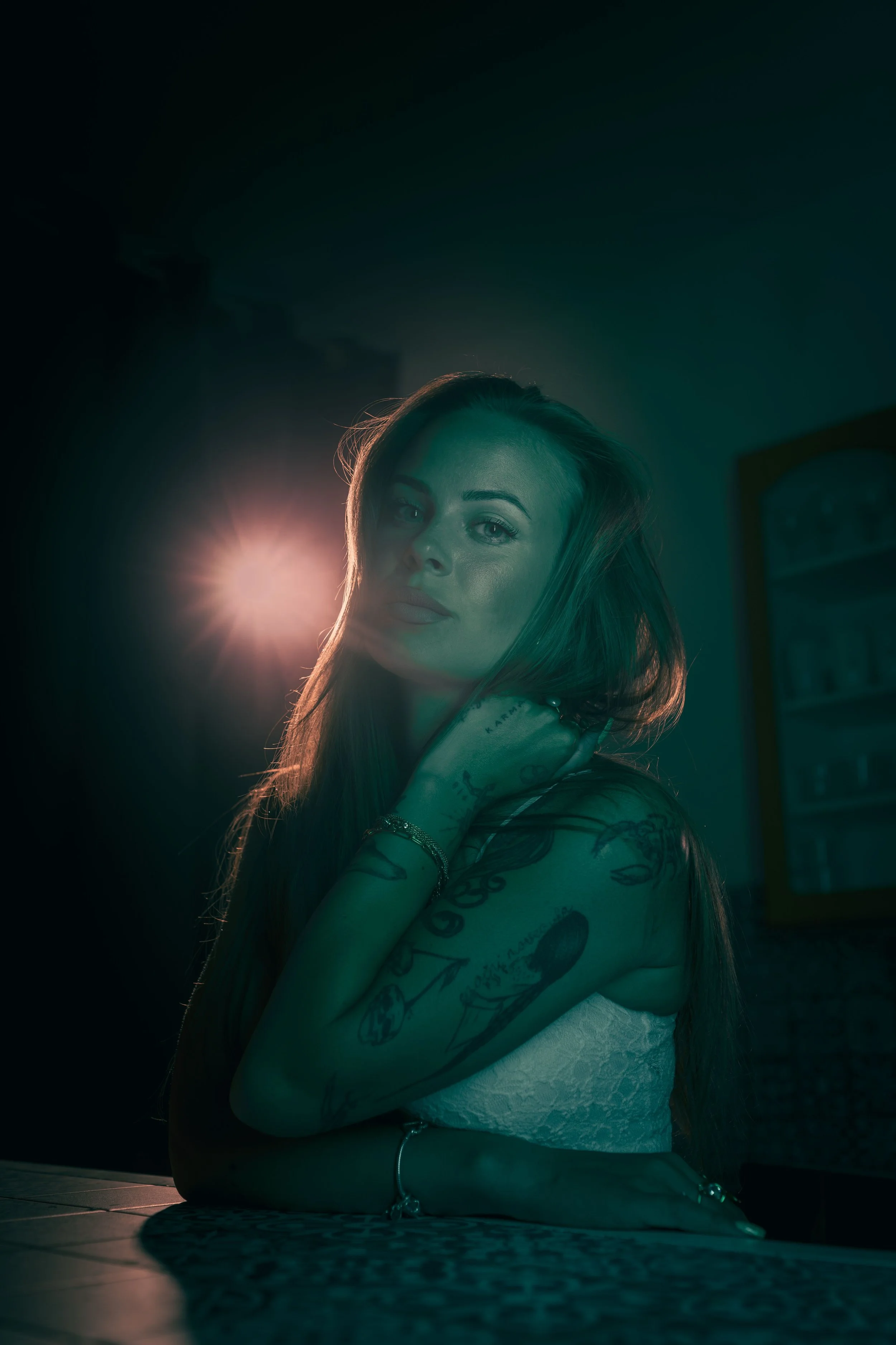 Fotografický portrét ženy s tetováním na pažích a ramenou, dlouhými vlasy a světlou pletí, sedící na vzorovaném povrchu s hlavou opřenou o ruku, osvětlená jasným světlem za ní.
