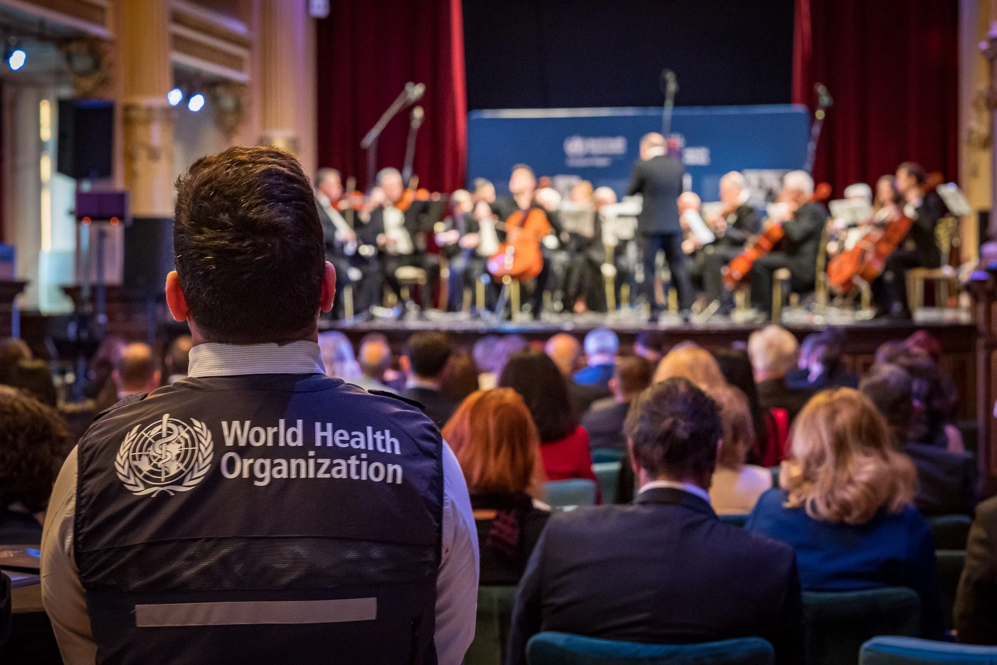 A person wearing a World Health Organization vest attending a concert by an orchestra with a conductor on stage and an audience in a large theater.