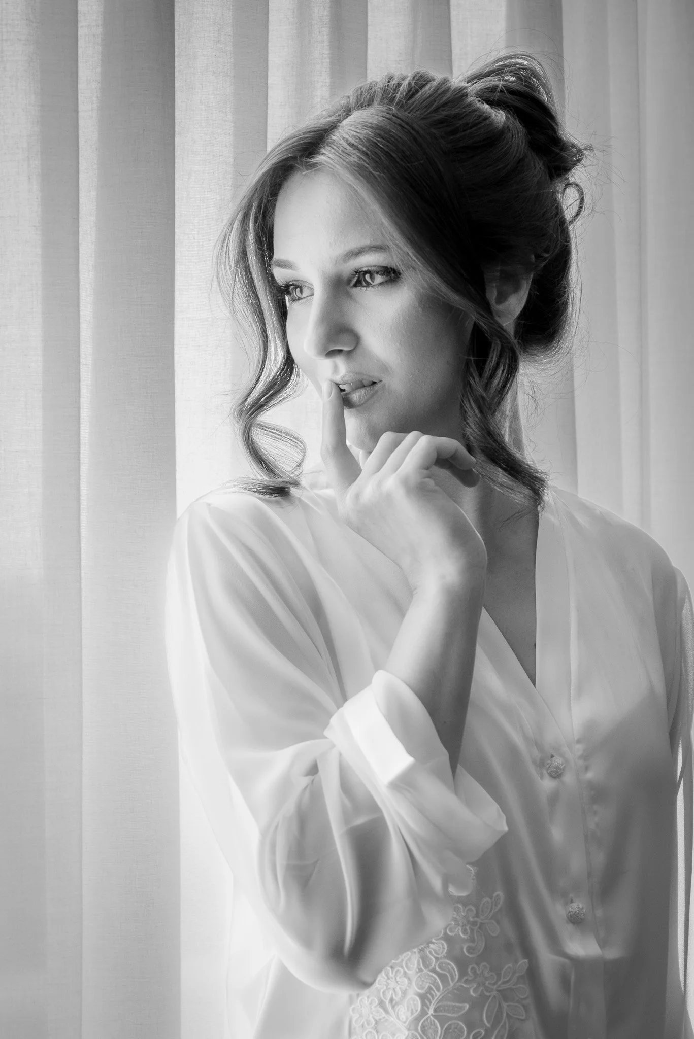 Žena s upravenými vlasy v saténové košili stojí u průsvitné záclony a má prst na rtech v zamyšlené póze.