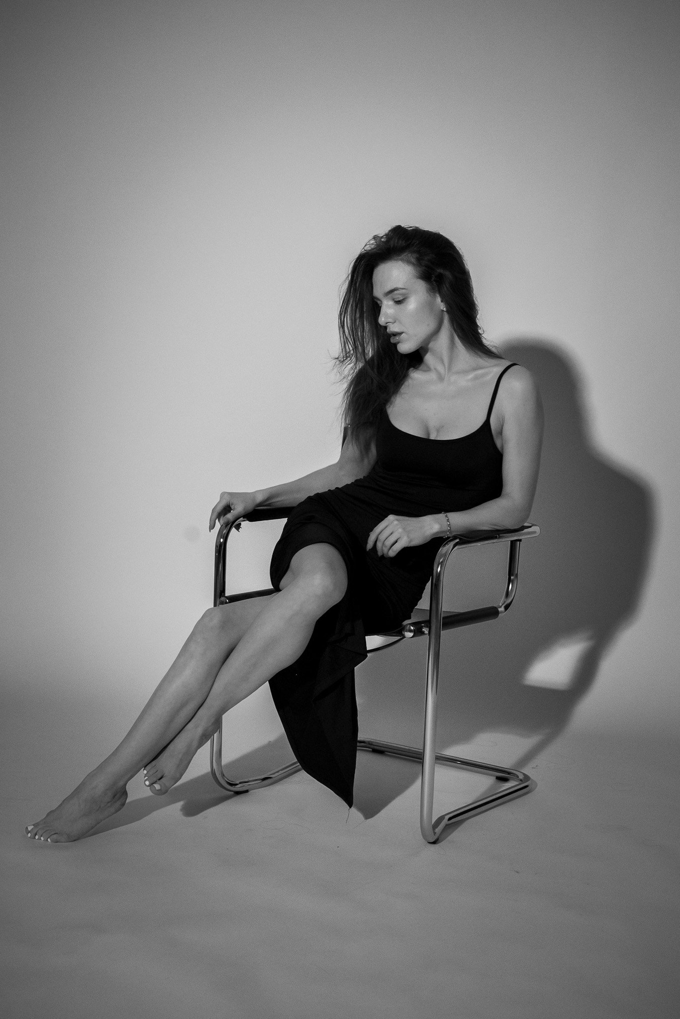 Black and white photo of a woman sitting on a chair, wearing a black dress, with crossed legs and looking down.