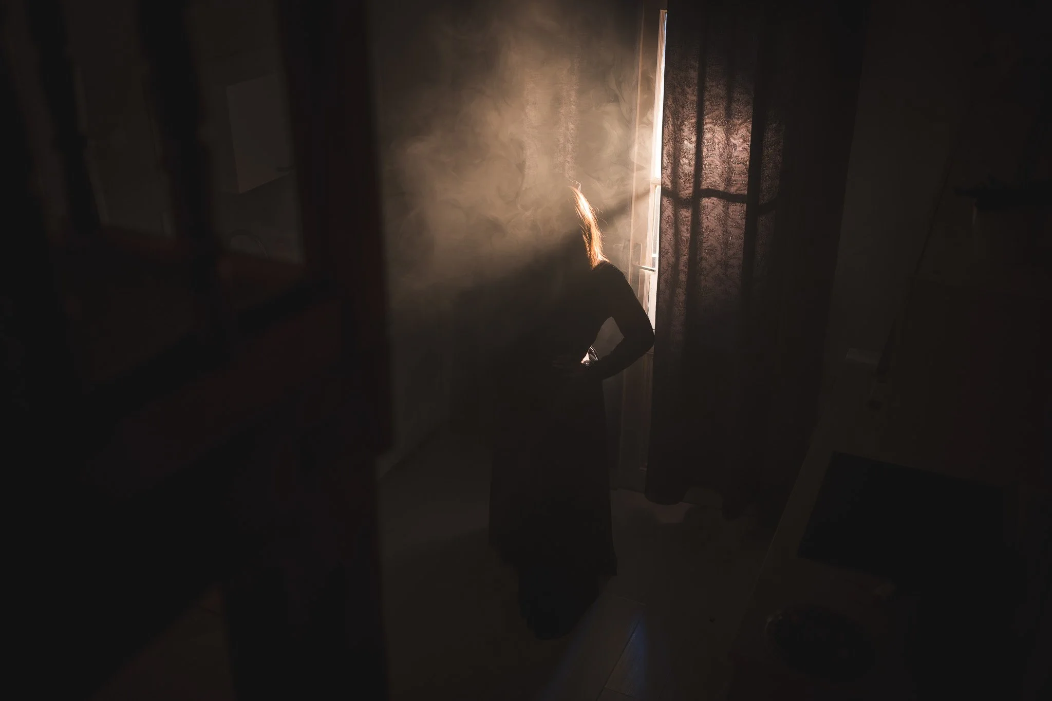 A woman with blonde hair standing in a dimly lit room, with sunlight streaming through a partially open door with curtains.