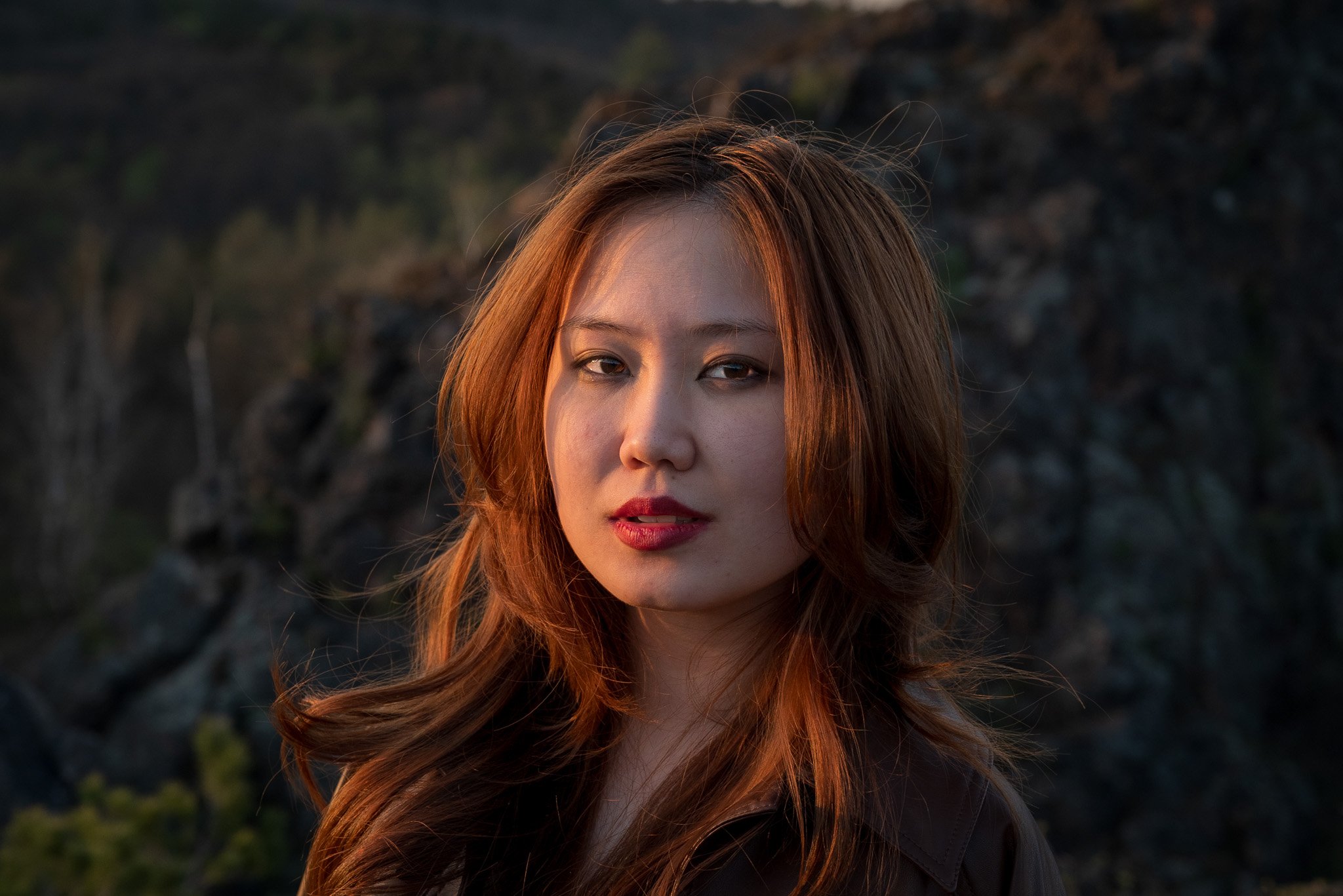 Woman with red hair standing in front of a rocky landscape, illuminated by sunset light.