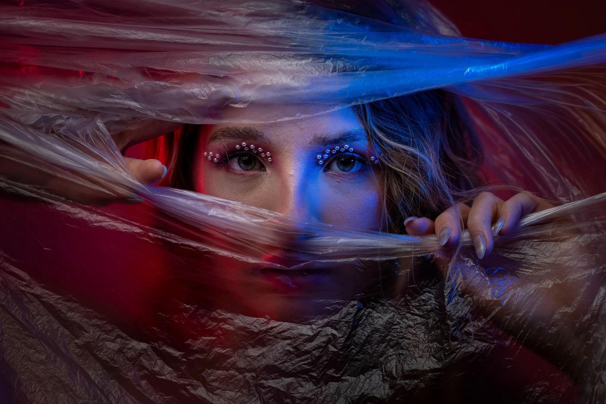 A woman peering through a plastic sheet, with metallic makeup, rhinestones around her eyes, and dramatic lighting in red and blue tones.