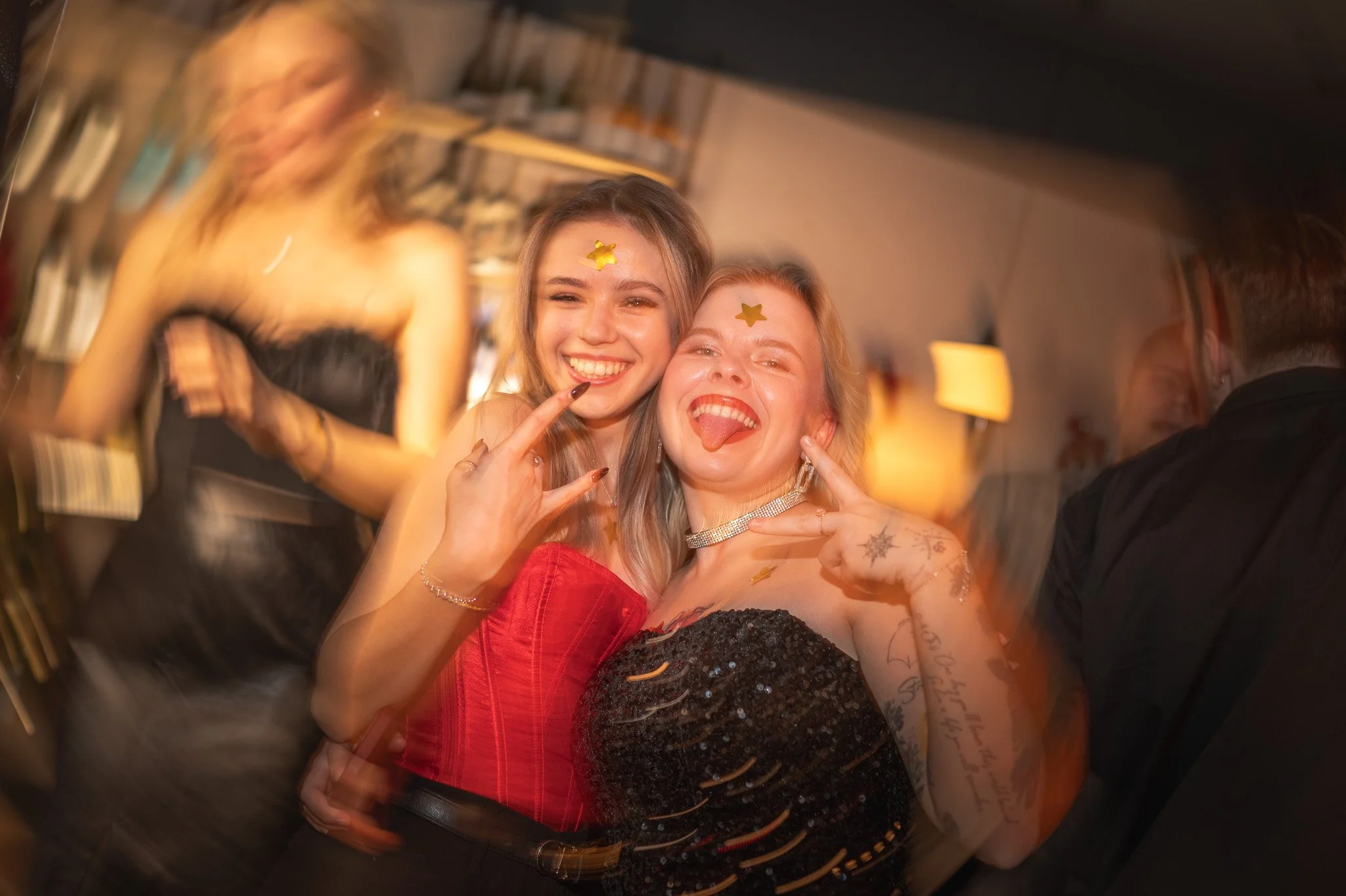 Two women with star stickers on their foreheads smiling and making peace signs at a lively party.