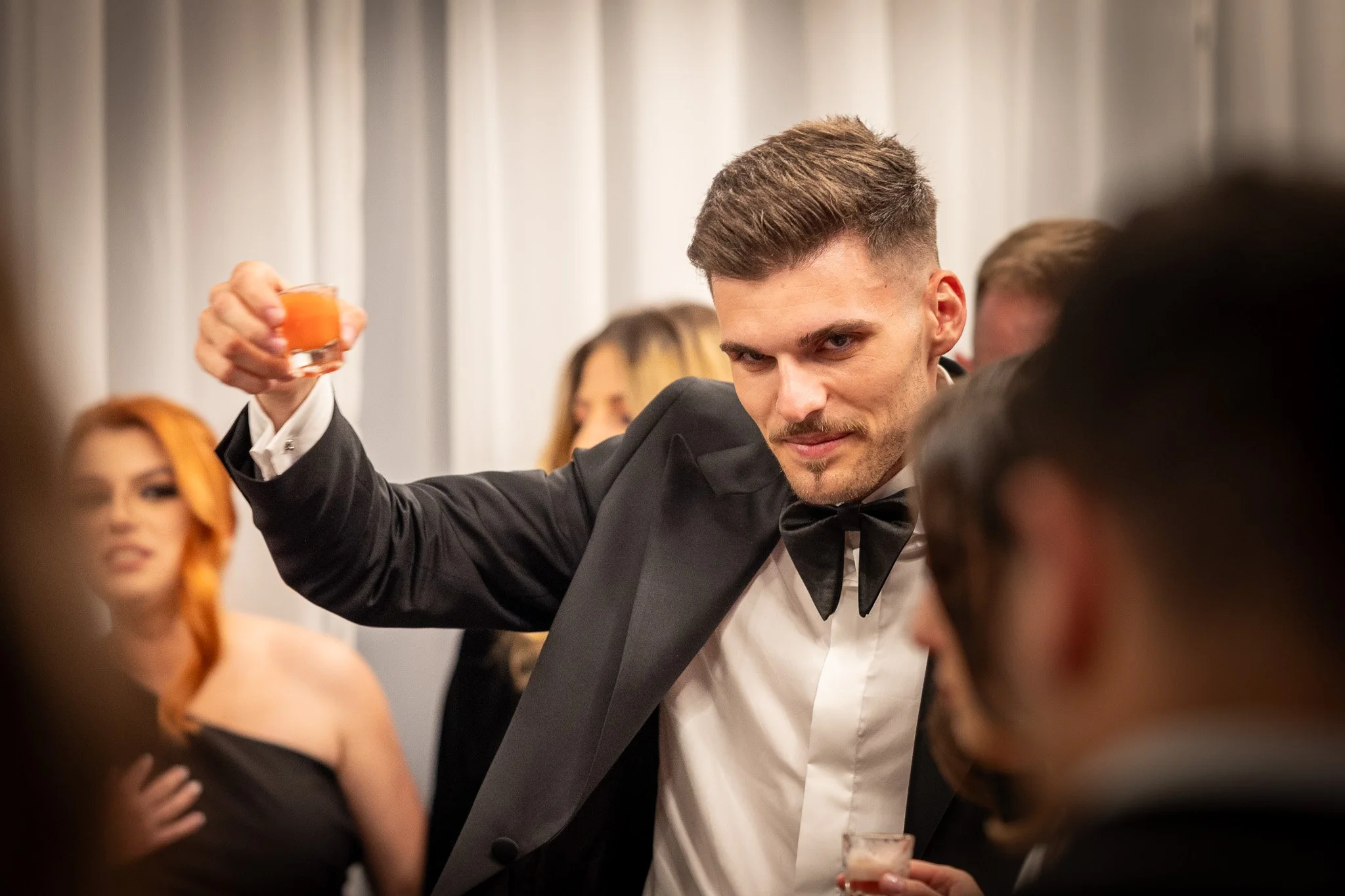 Man in a tuxedo raising a glass at a formal party with other guests in the background.