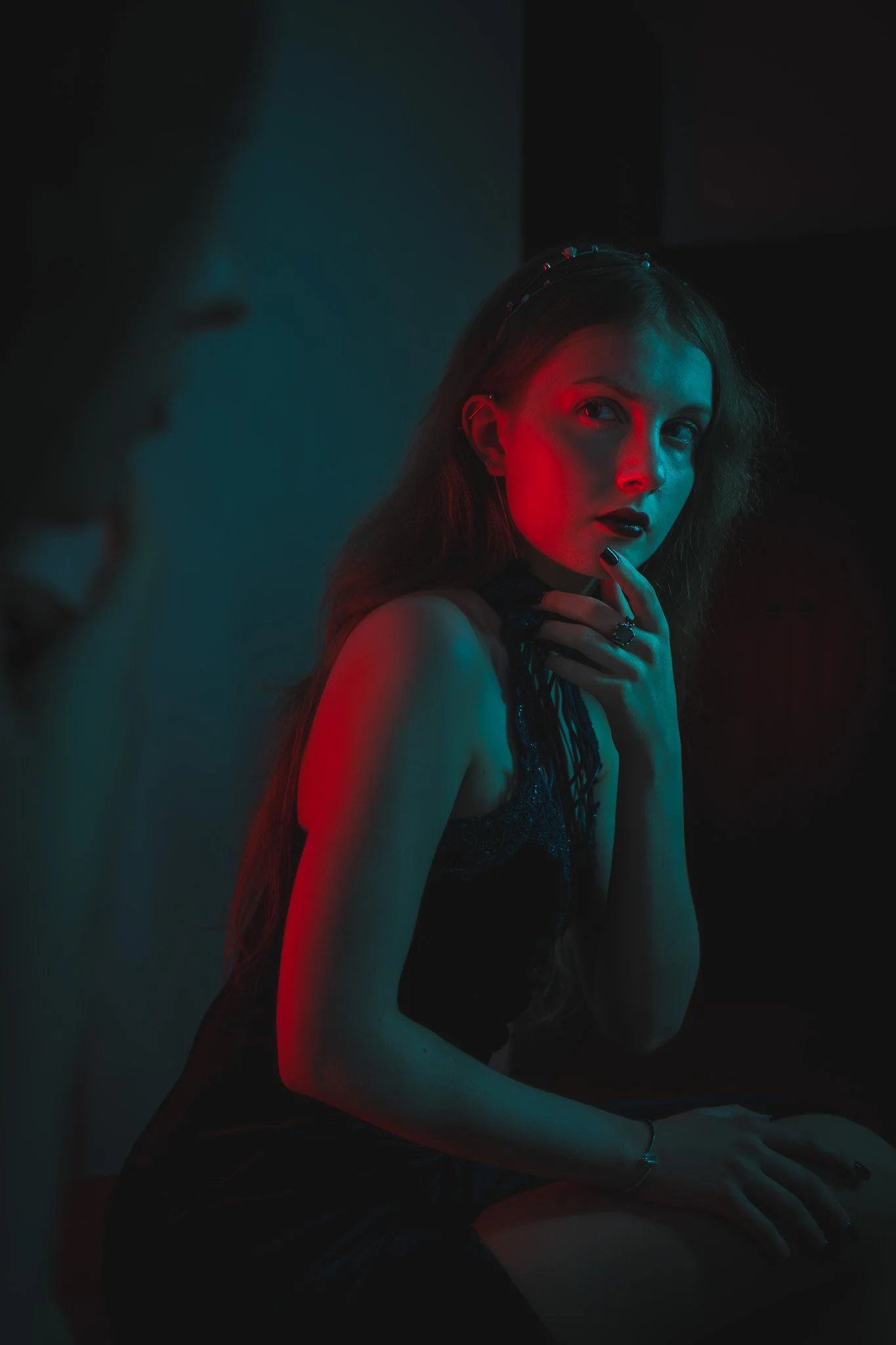 A woman with long hair and dark lipstick sitting in a dimly lit room illuminated by red and blue lights, wearing a black lace top and accessories, with a pensive expression.