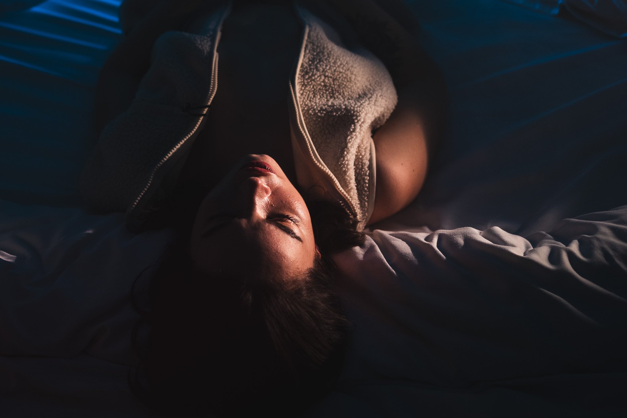 A woman lying on a bed in dim lighting, with her eyes closed, wearing a jacket and a black top.