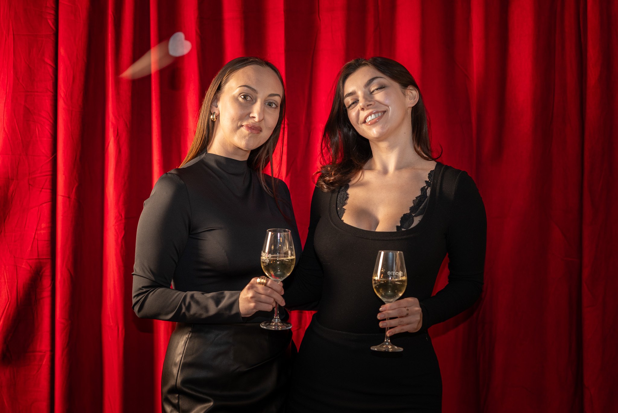 Two women in black dresses holding glasses of white wine in front of a red velvet curtain.