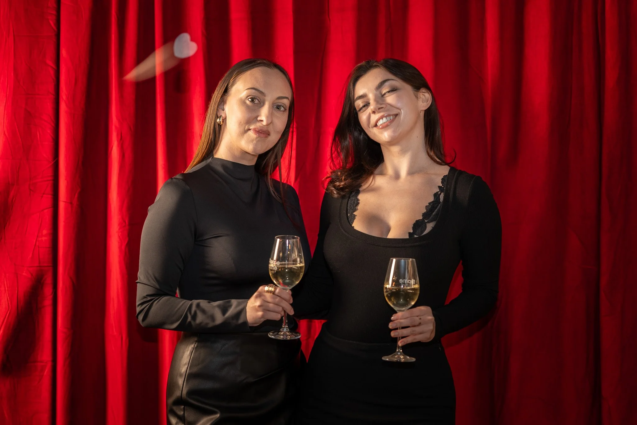 Two women in black dresses holding glasses of white wine in front of a red velvet curtain.