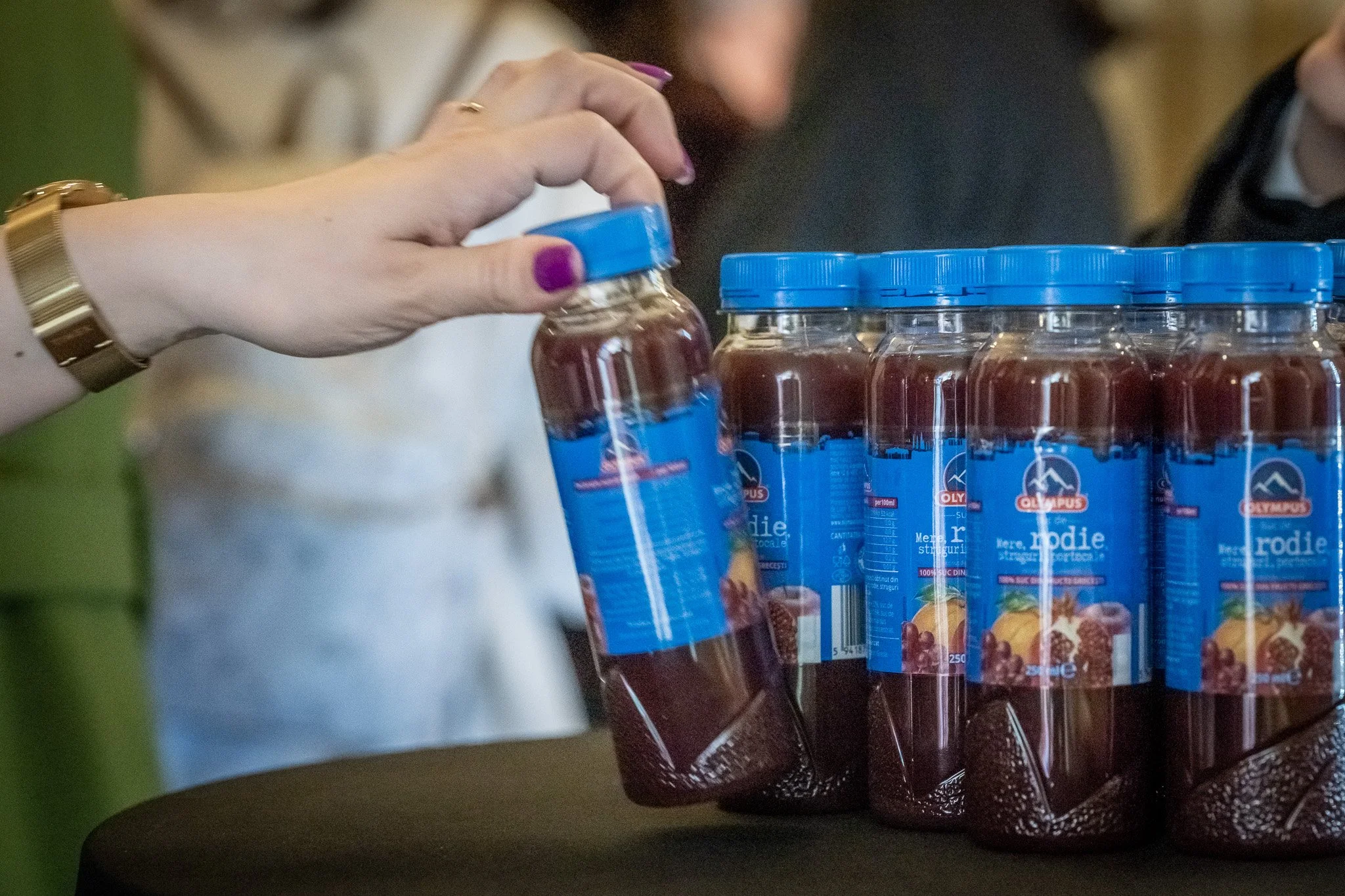 A person's hand, with purple nail polish and a gold watch, is placing a jar of red jelly with a blue label on a table. Several similar jars of red jelly with blue labels are already on the table.